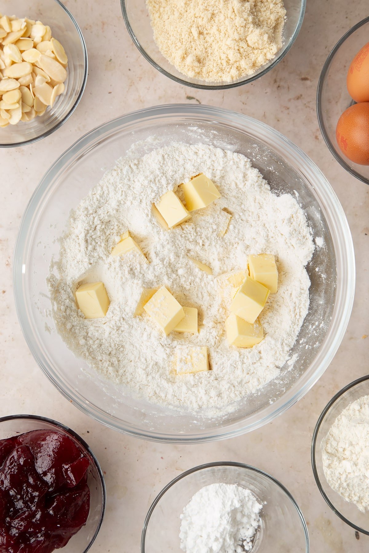 Flour, icing sugar and butter in a bowl. Ingredients to make a Bakewell tray bake surround the bowl.