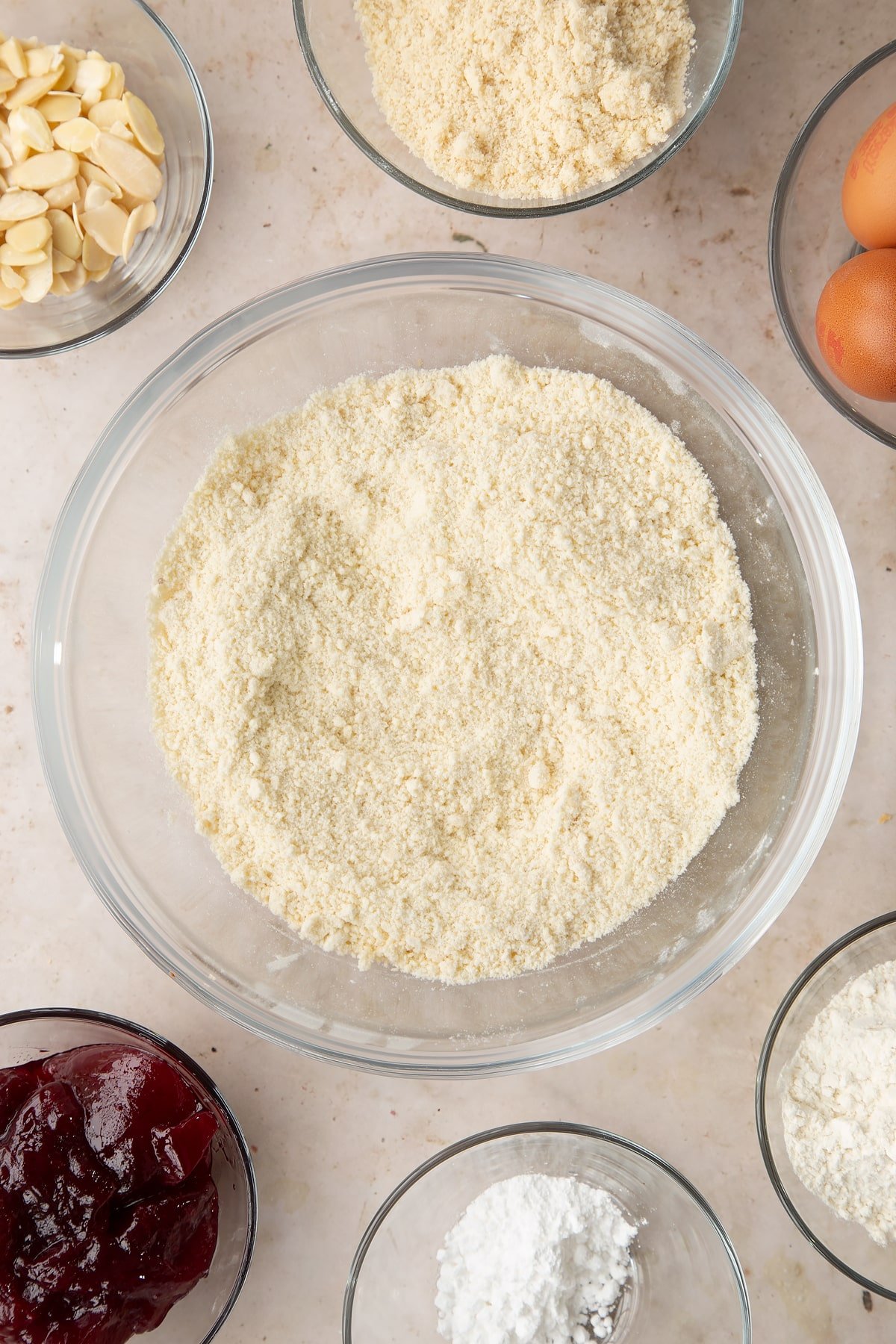 Flour, icing sugar and butter rubbed together in a bowl. Ingredients to make a Bakewell tray bake surround the bowl.