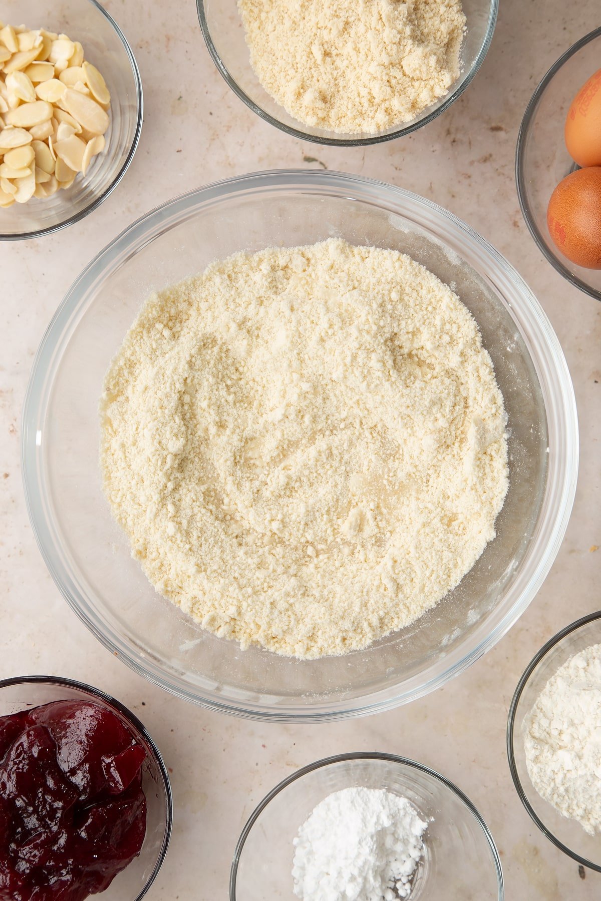 Flour, icing sugar and butter rubbed together in a bowl with a little water on top. Ingredients to make a Bakewell tray bake surround the bowl.