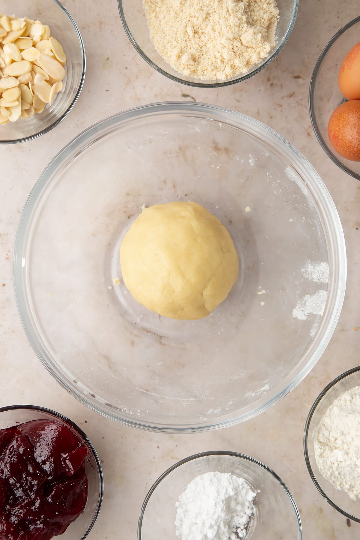 A ball of pastry in a bowl. Ingredients to make a Bakewell tray bake surround the bowl.