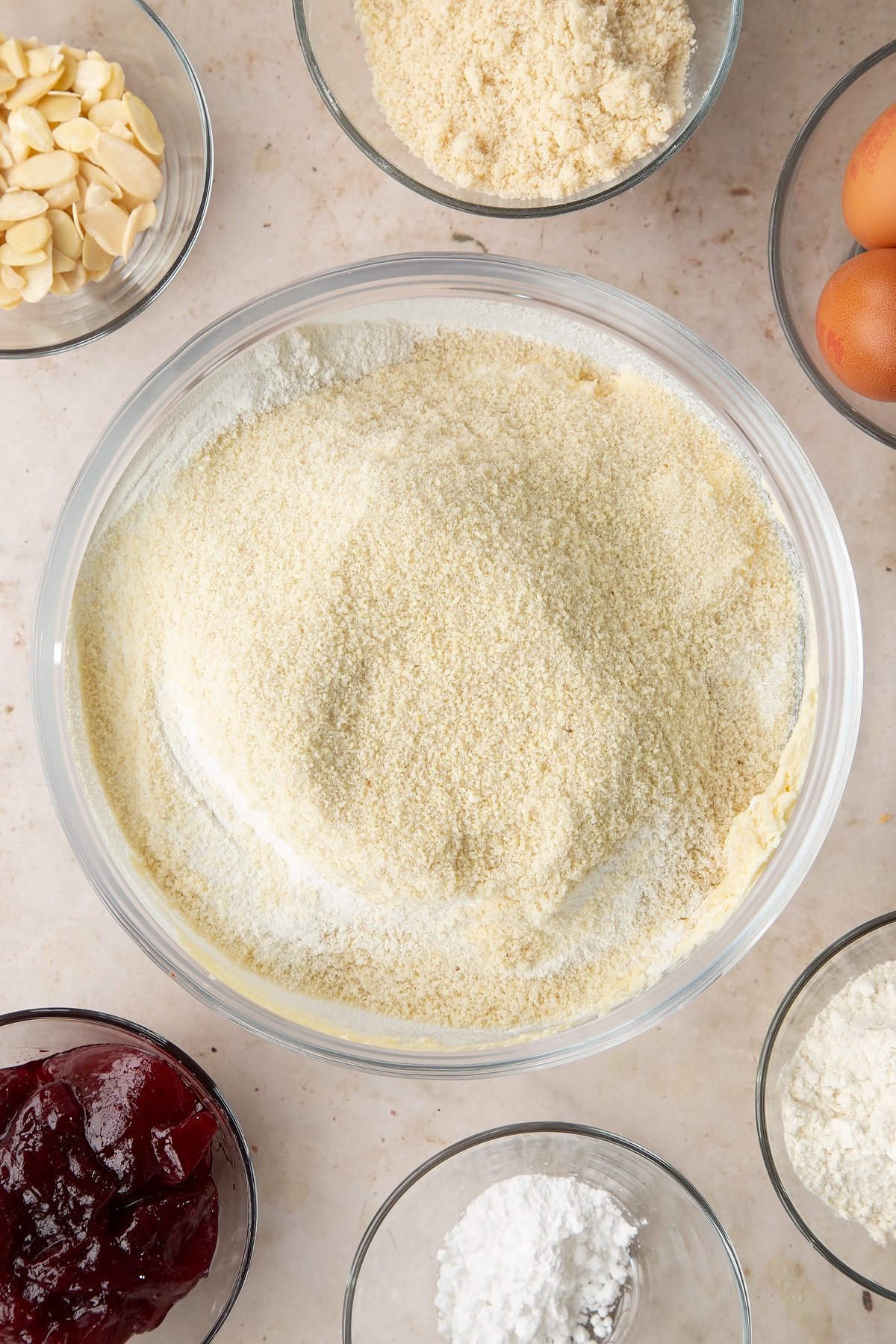 Butter, sugar and eggs creamed together in a bowl with almonds and flour on top. Ingredients to make a Bakewell tray bake surround the bowl.