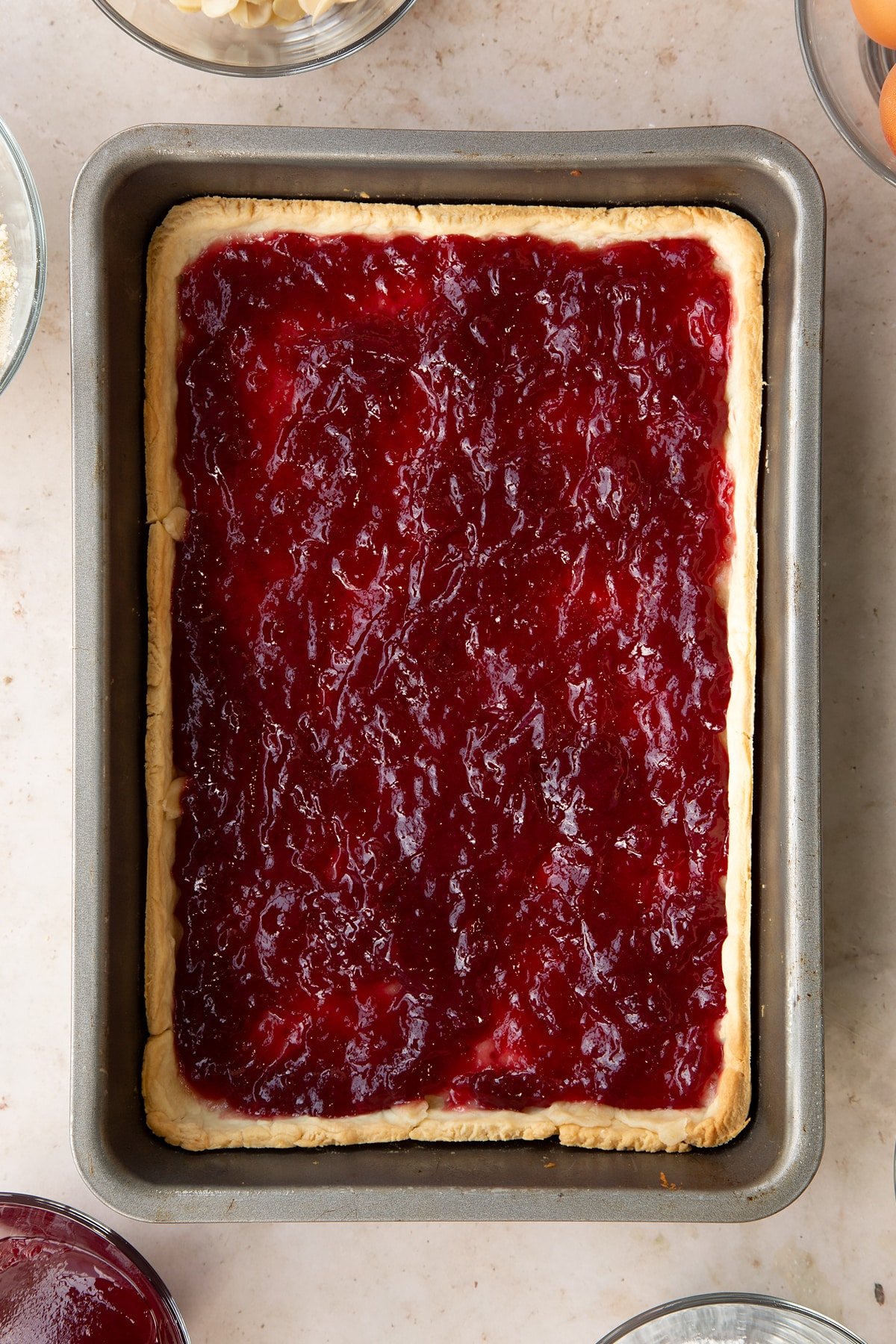 Part-cooked pastry in a rectangular tin. It has been spread with jam.