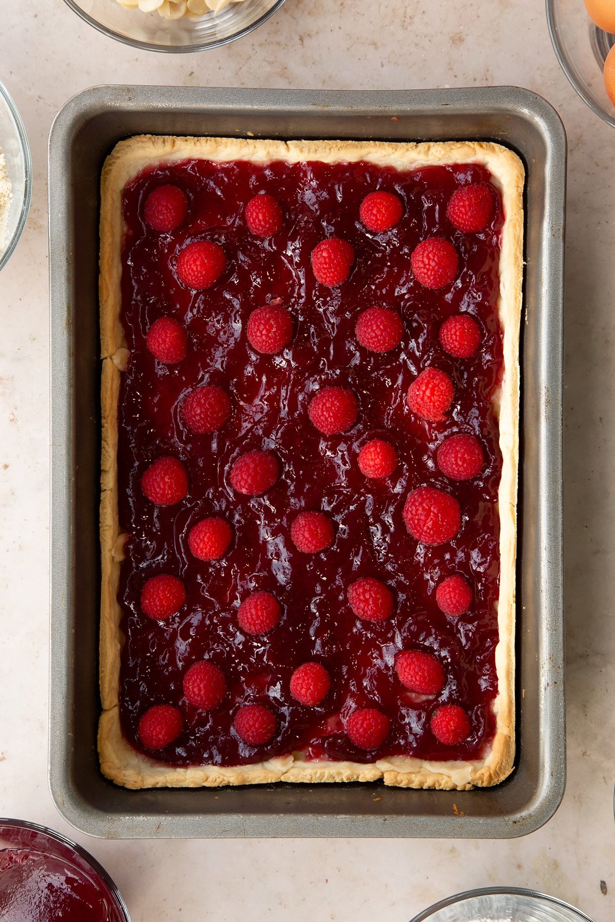 Part-cooked pastry in a rectangular tin. It has been spread with jam and dotted with raspberries.