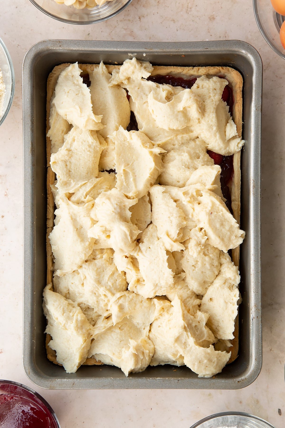 Bakewell tray bake batter in a tin in dollops, topping pastry and jam, shown from above.