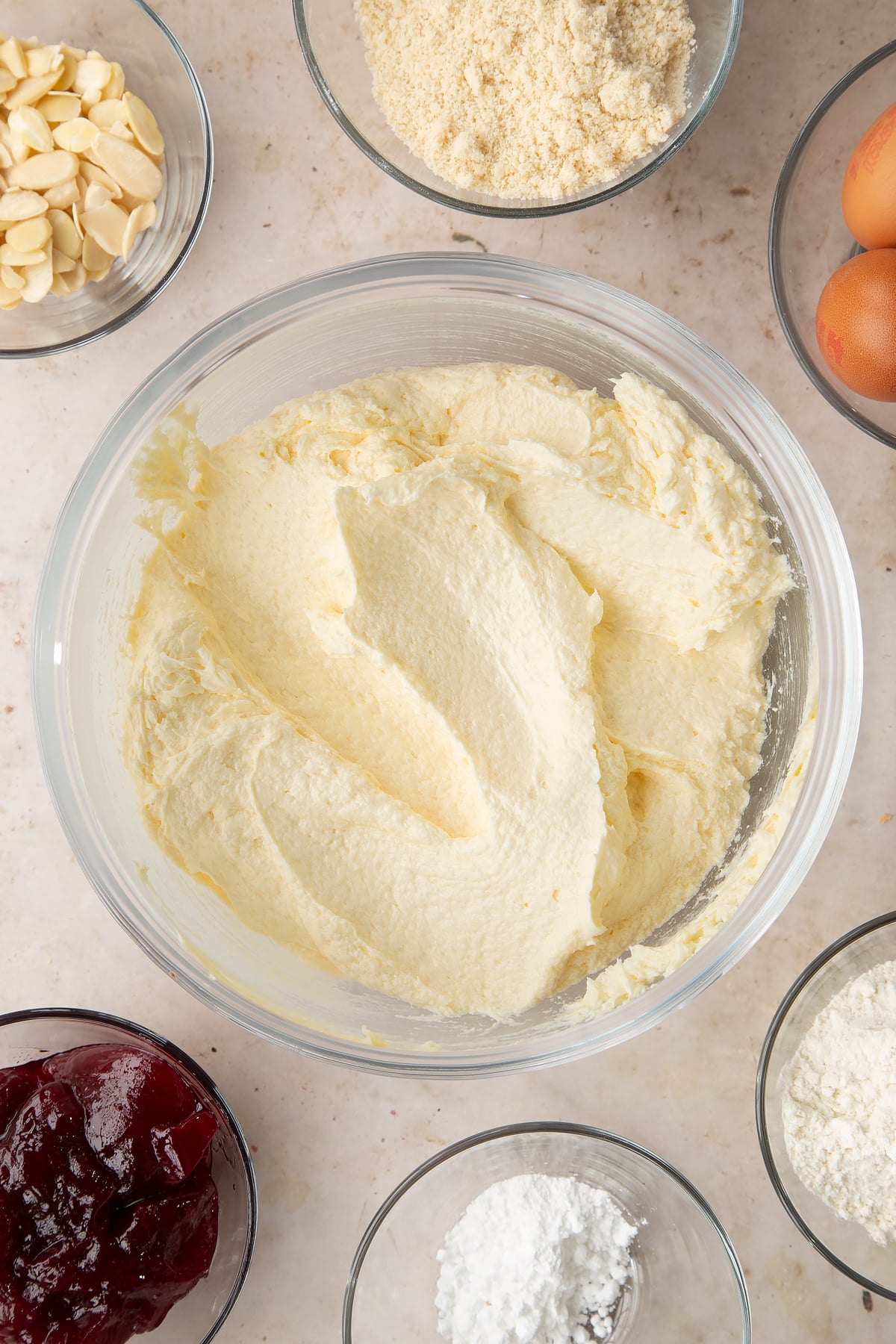 Butter, sugar and eggs creamed together in a bowl. Ingredients to make a Bakewell tray bake surround the bowl.