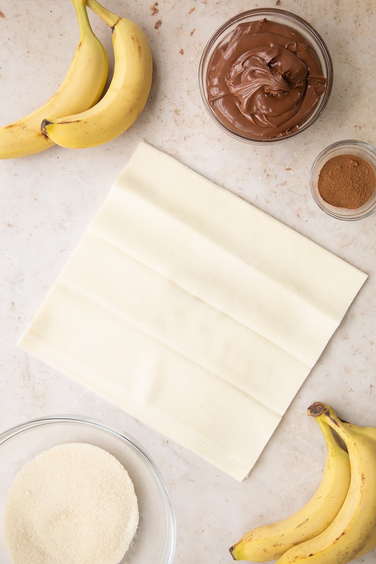 Overhead shot of the filo pastry rolled out on a table with some of the ingredients required for the banana chocolate spring rolls on the side.