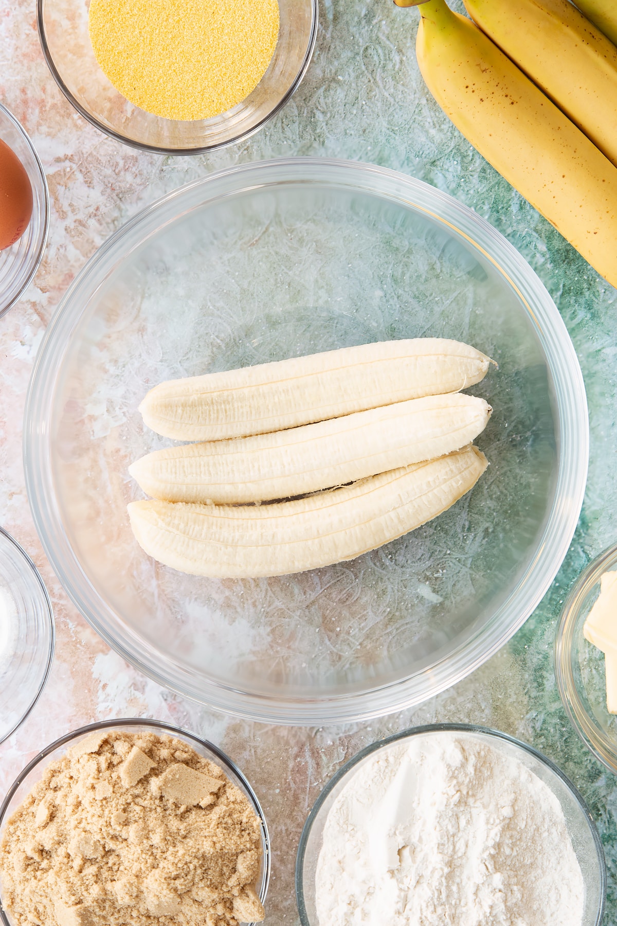 Bananas in a bowl. Ingredients to make banana cornmeal muffin surround the bowl.