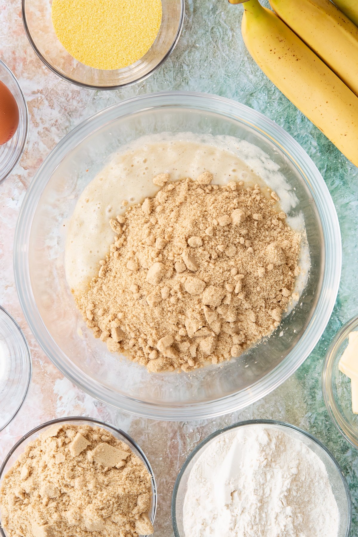 Mashed bananas in a bowl with sugar on top. Ingredients to make banana cornmeal muffin surround the bowl.