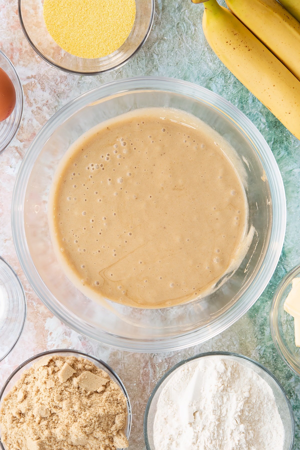 Mashed bananas and sugar mixed together in a bowl. Ingredients to make banana cornmeal muffin surround the bowl.