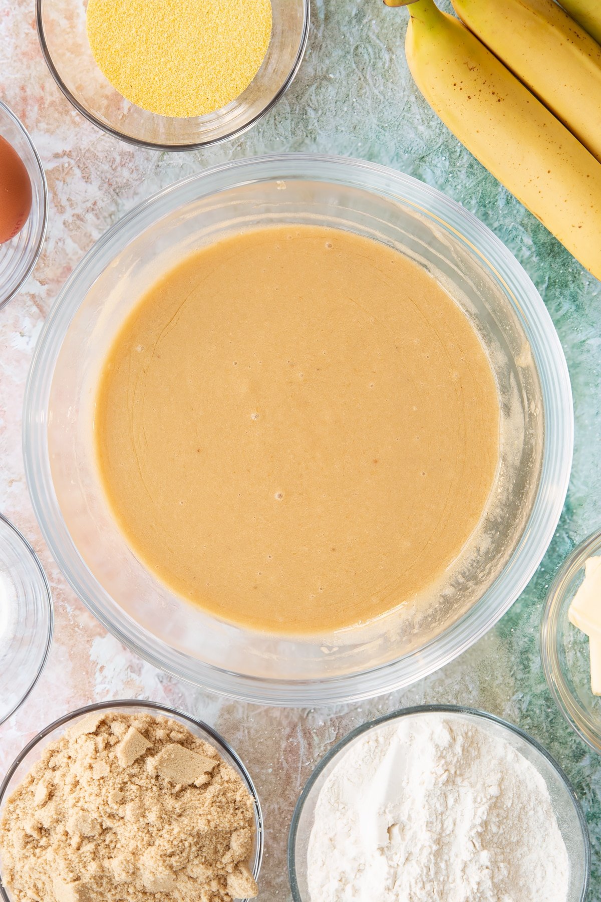 Mashed bananas, melted butter and sugar mixed together in a bowl. Ingredients to make banana cornmeal muffin surround the bowl.