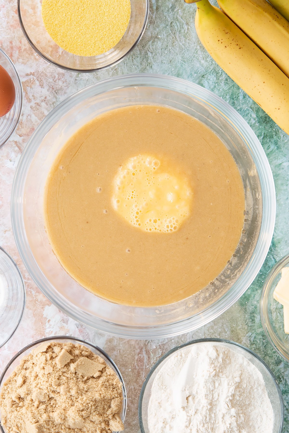 Mashed bananas, melted butter and sugar mixed together in a bowl with beaten eggs on top. Ingredients to make banana cornmeal muffin surround the bowl.