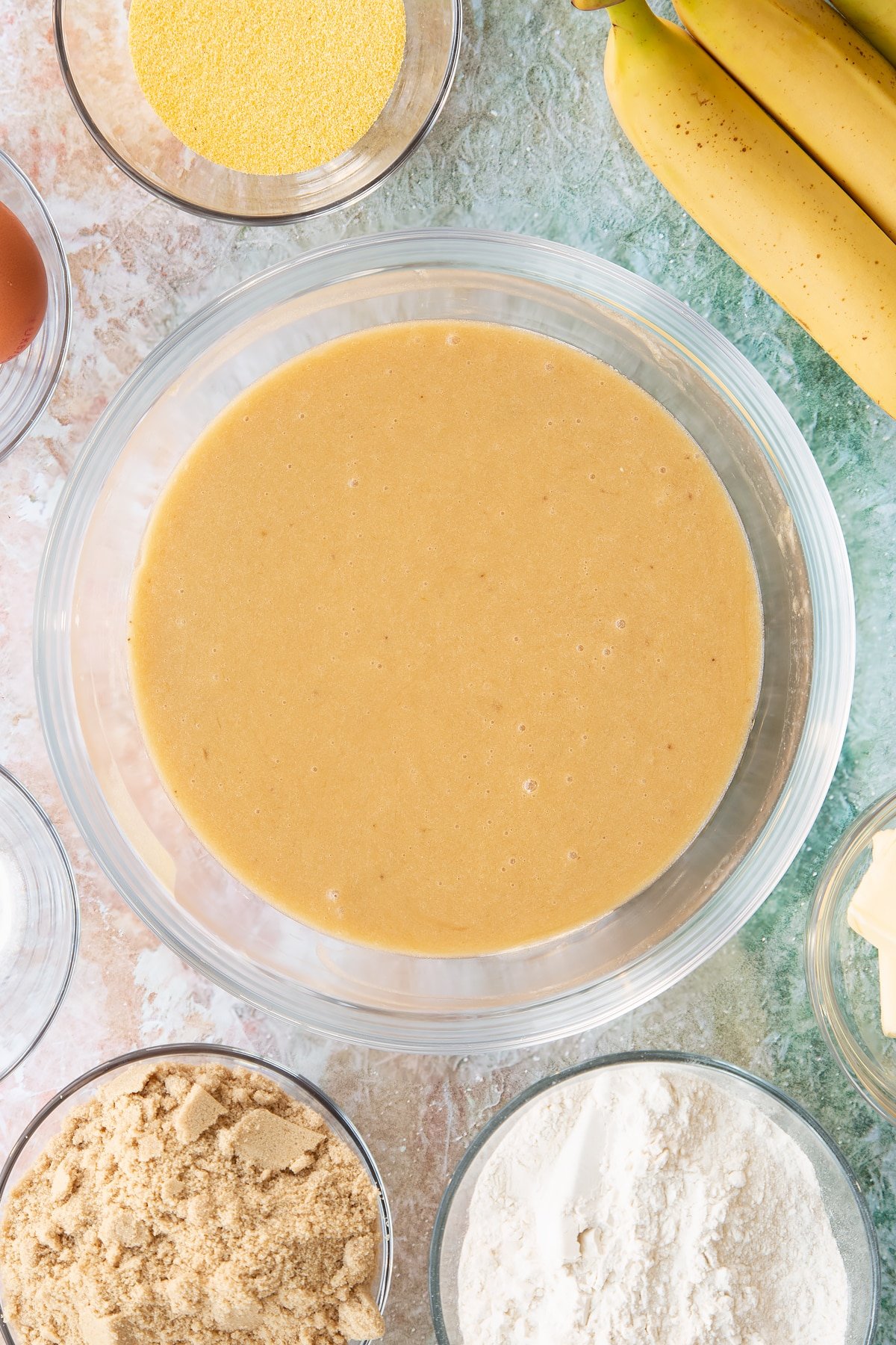 Mashed bananas, melted butter, eggs and sugar mixed together in a bowl. Ingredients to make banana cornmeal muffin surround the bowl.