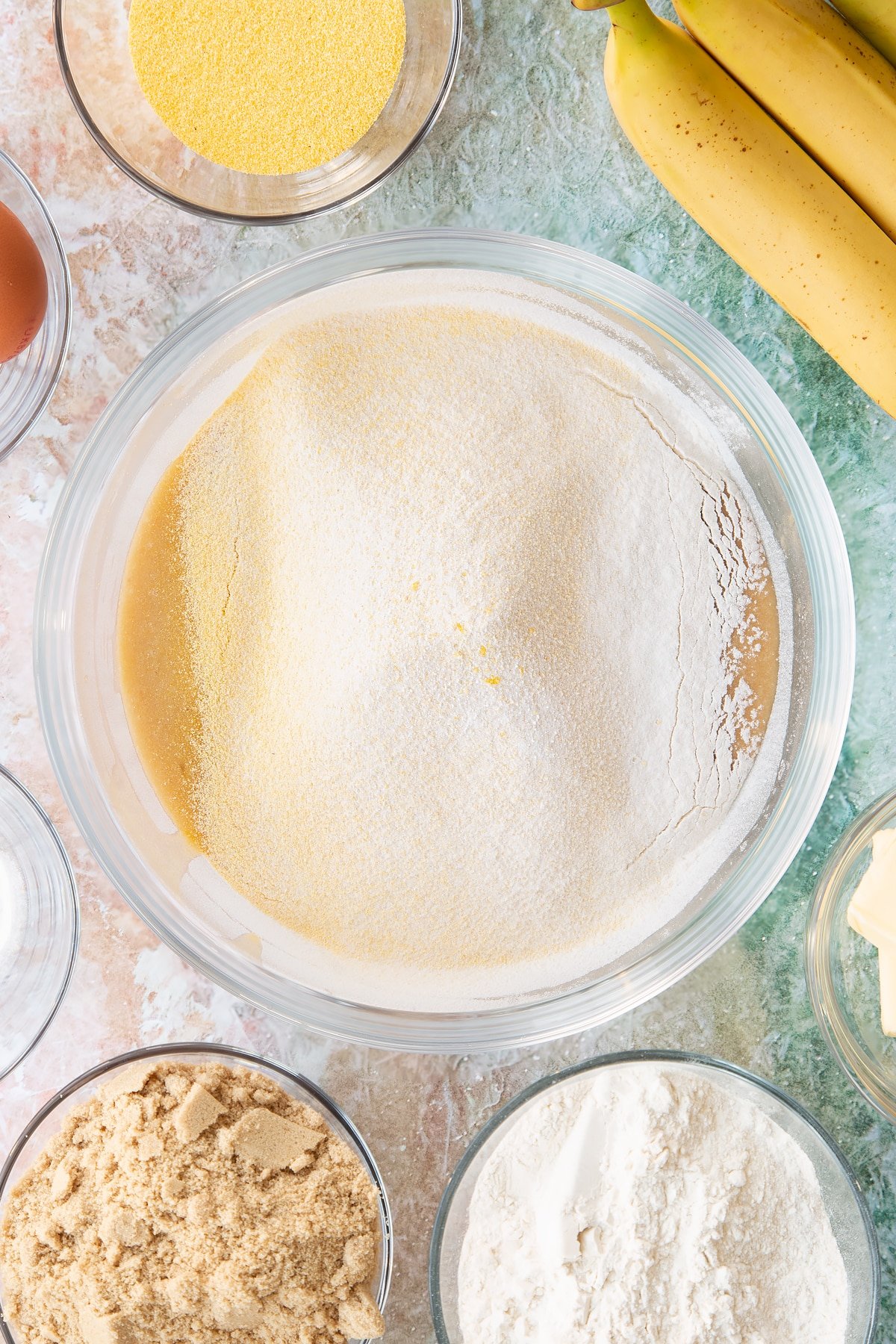 Mashed bananas, melted butter, eggs and sugar mixed together in a bowl with flour and cornmeal on top. Ingredients to make banana cornmeal muffin surround the bowl.