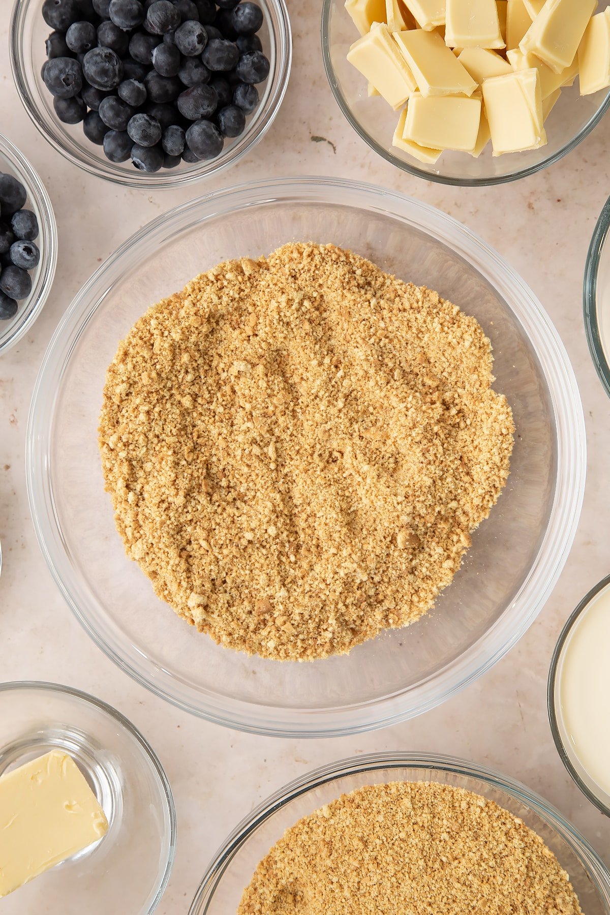 Crushed biscuits in a bowl. Ingredients to make blueberry cheesecake pie surround the bowl.