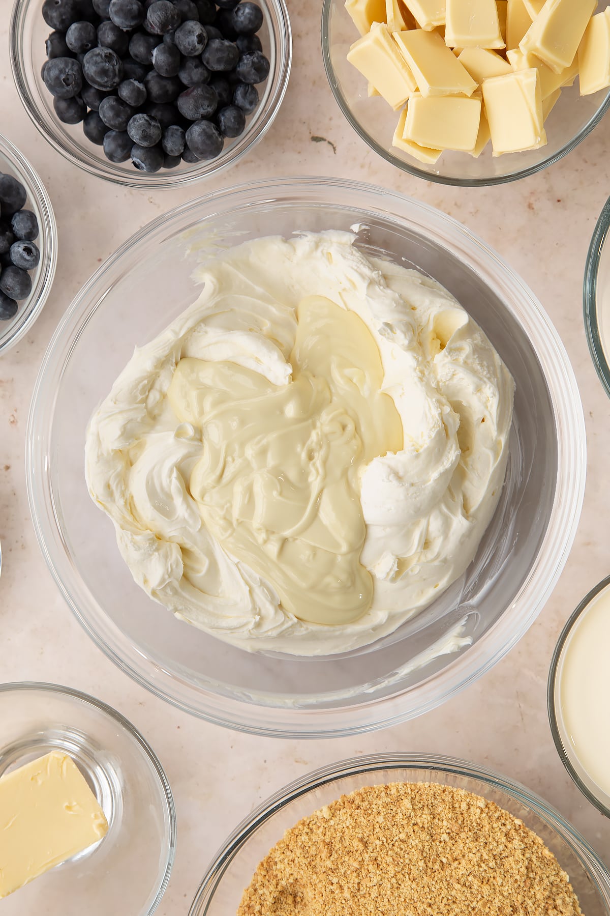 Whipped cream mixed with cream cheese, sugar and vanilla in a bowl with white chocolate on top. Ingredients to make blueberry cheesecake pie surround the bowl.