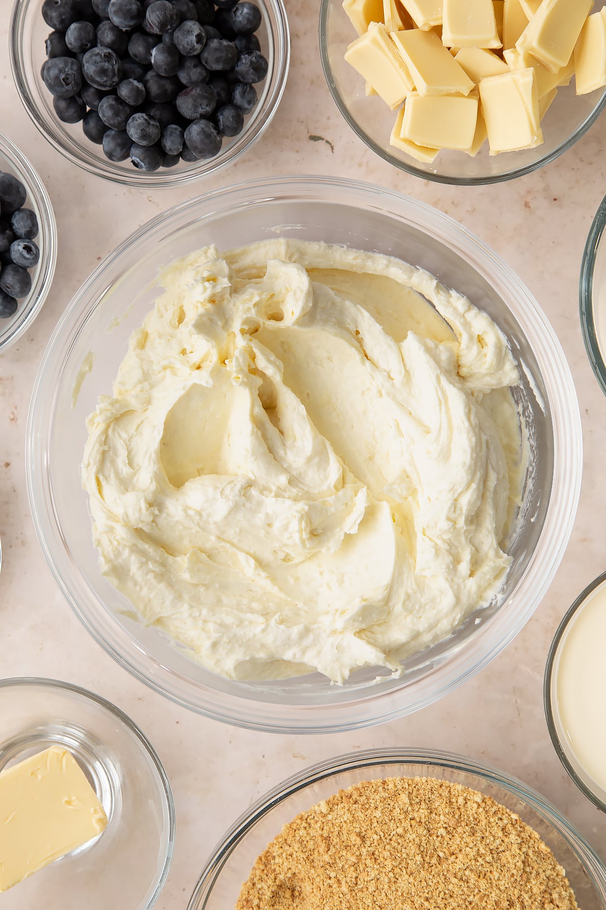 Cheesecake filling in a bowl. Ingredients to make blueberry cheesecake pie surround the bowl.