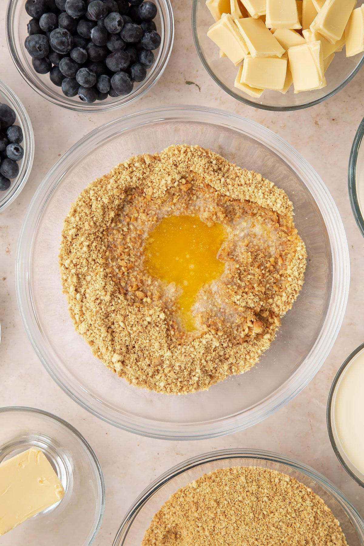 Crushed biscuits and melted butter in a bowl. Ingredients to make blueberry cheesecake pie surround the bowl.