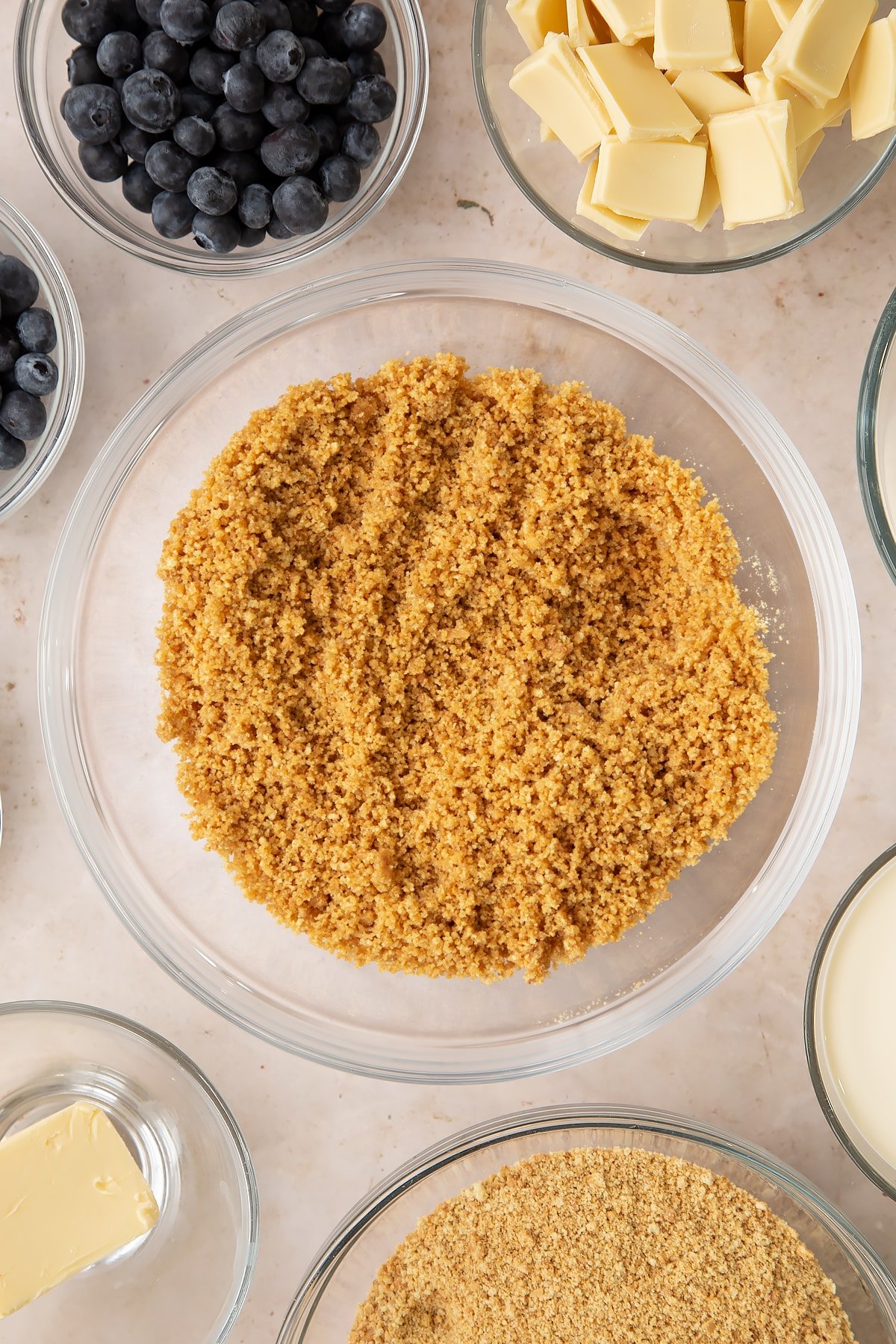 Crushed biscuits and melted butter mixed together in a bowl. Ingredients to make blueberry cheesecake pie surround the bowl.