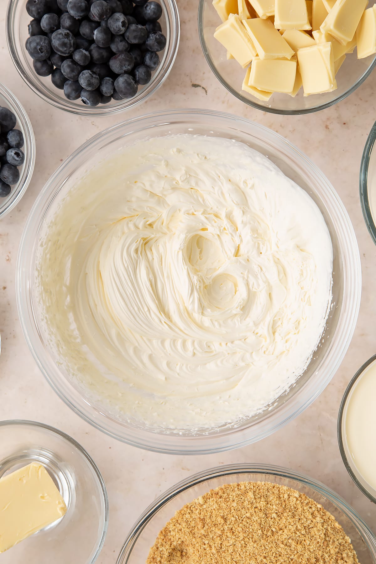 Whipped cream in a bowl. Ingredients to make blueberry cheesecake pie surround the bowl.