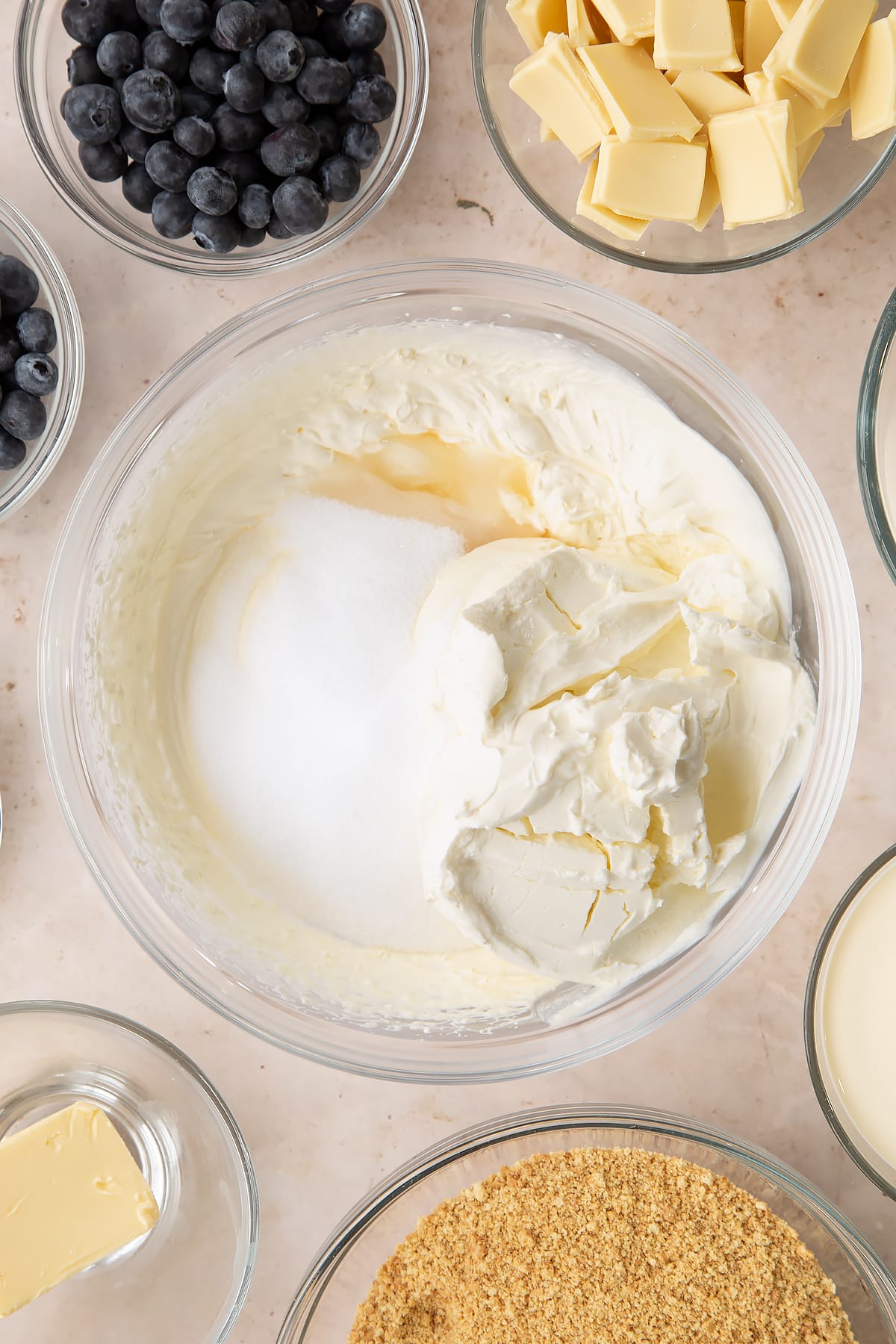 Whipped cream in a bowl with cream cheese, sugar and vanilla. Ingredients to make blueberry cheesecake pie surround the bowl.