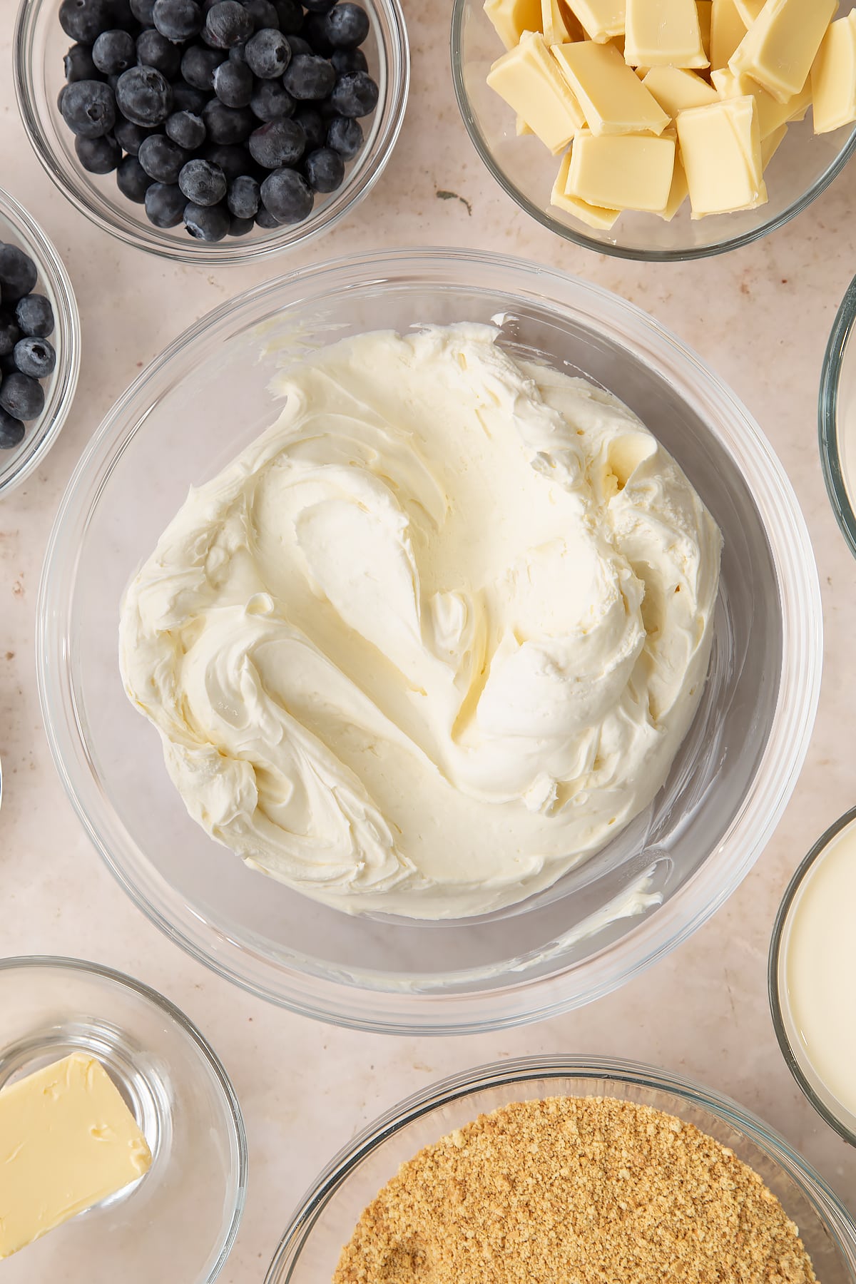 Whipped cream mixed with cream cheese, sugar and vanilla in a bowl. Ingredients to make blueberry cheesecake pie surround the bowl.