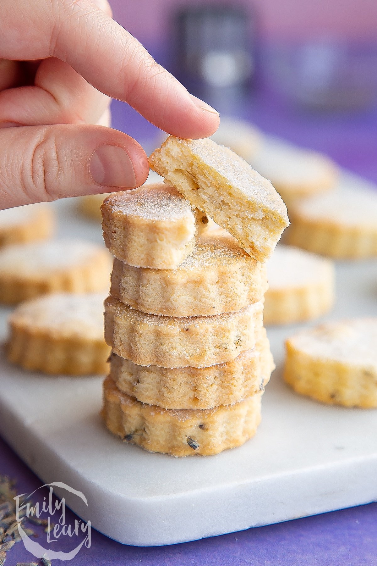 Lavender shortbread cookies in a stack on a marble board. The top one is broken in half and a hand reaches for it.