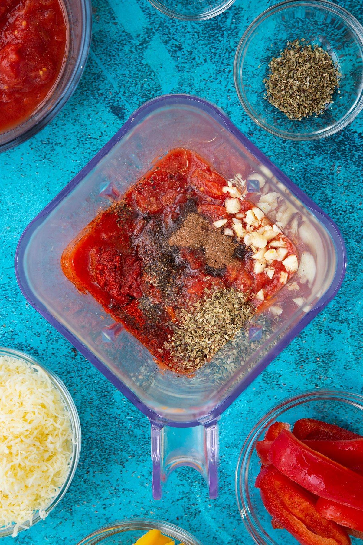 Tinned tomatoes, tomato puree, olive oil, vinegar and spices in a blender. Ingredients to make Quorn mince enchiladas surround the blender.