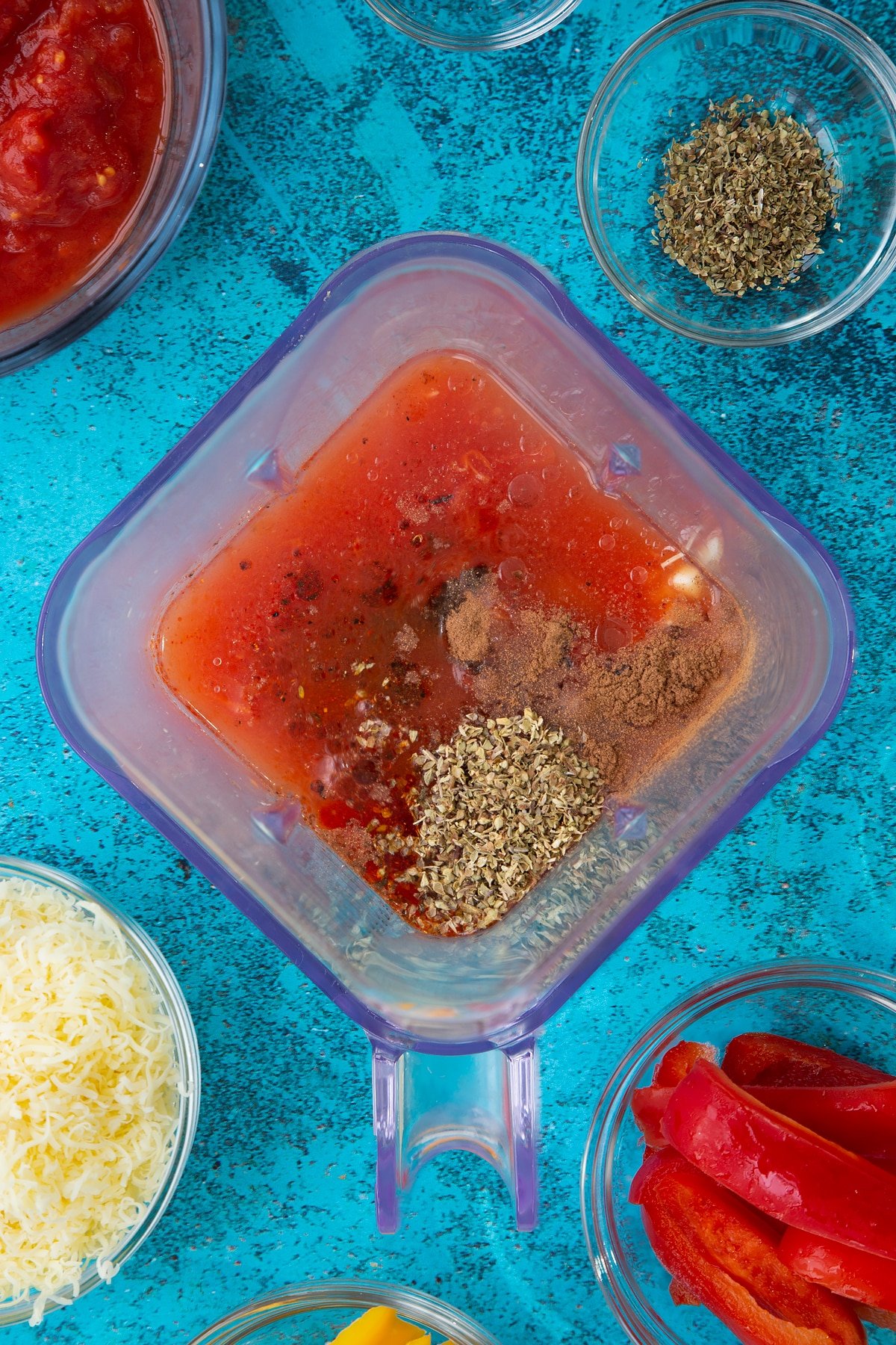 Water, tinned tomatoes, tomato puree, olive oil, vinegar and spices in a blender. Ingredients to make Quorn mince enchiladas surround the blender.
