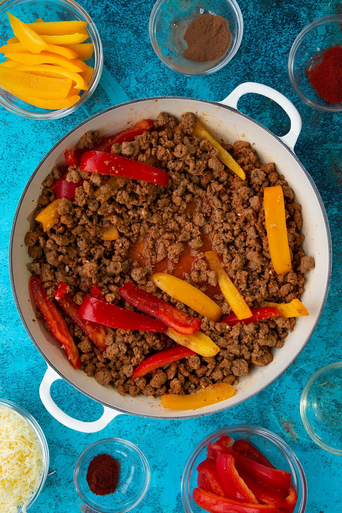 Quorn mince and peppers fried in garlic and spices with a splash of water in a large white pan. Ingredients to make Quorn mince enchiladas surround the pan.