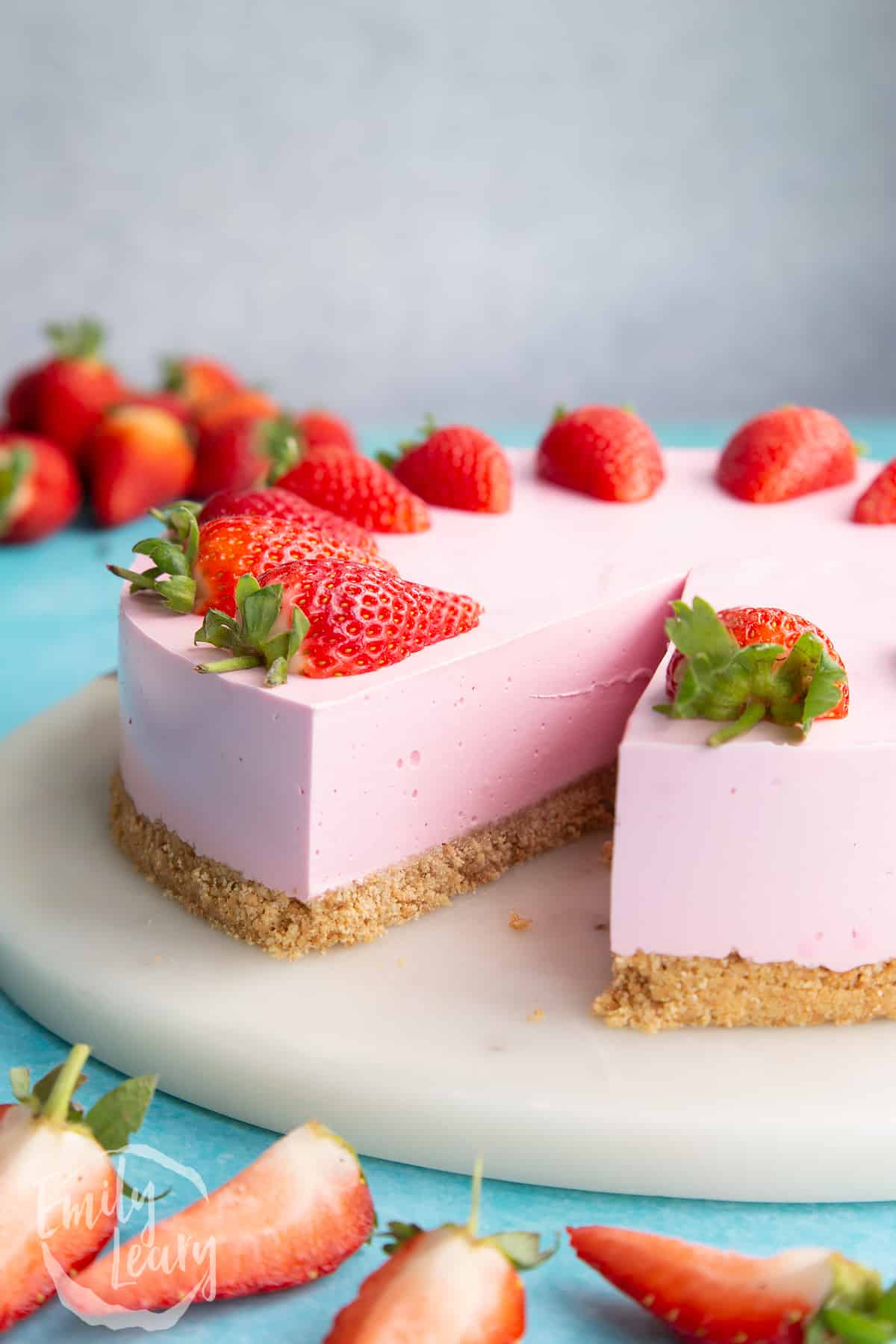 A pink jello cheesecake topped with a strawberry halves on a white marble board. A slice has been taken.