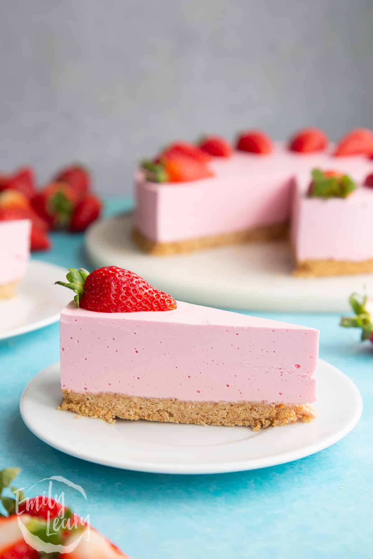 A slice of pink jello cheesecake on a white plate. It is topped with a strawberry half.
