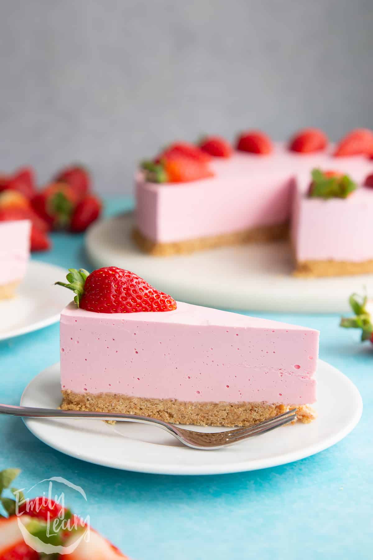 A slice of pink jello cheesecake on a white plate. It is topped with a strawberry half. A fork rests on the plate.