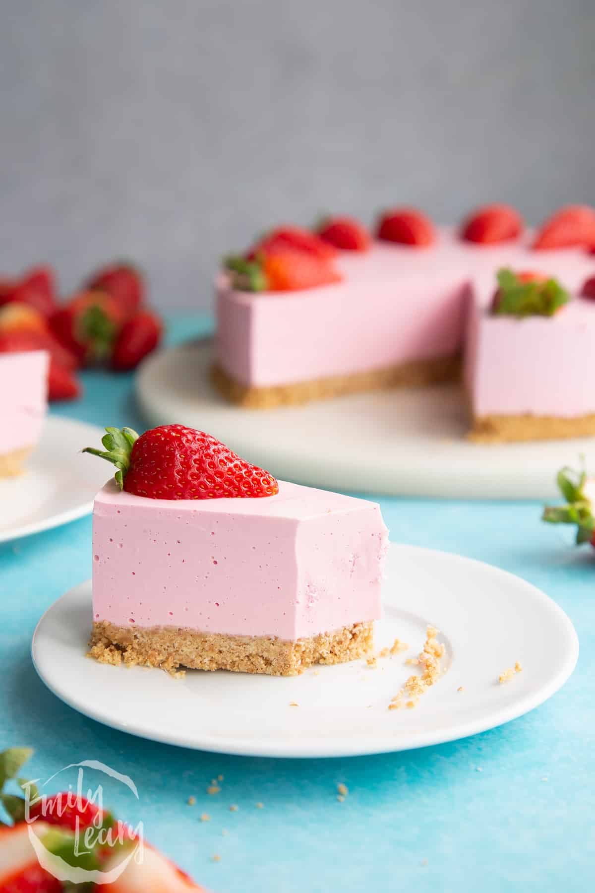 A slice of pink jello cheesecake on a white plate. It is topped with a strawberry half. Part has been eaten.