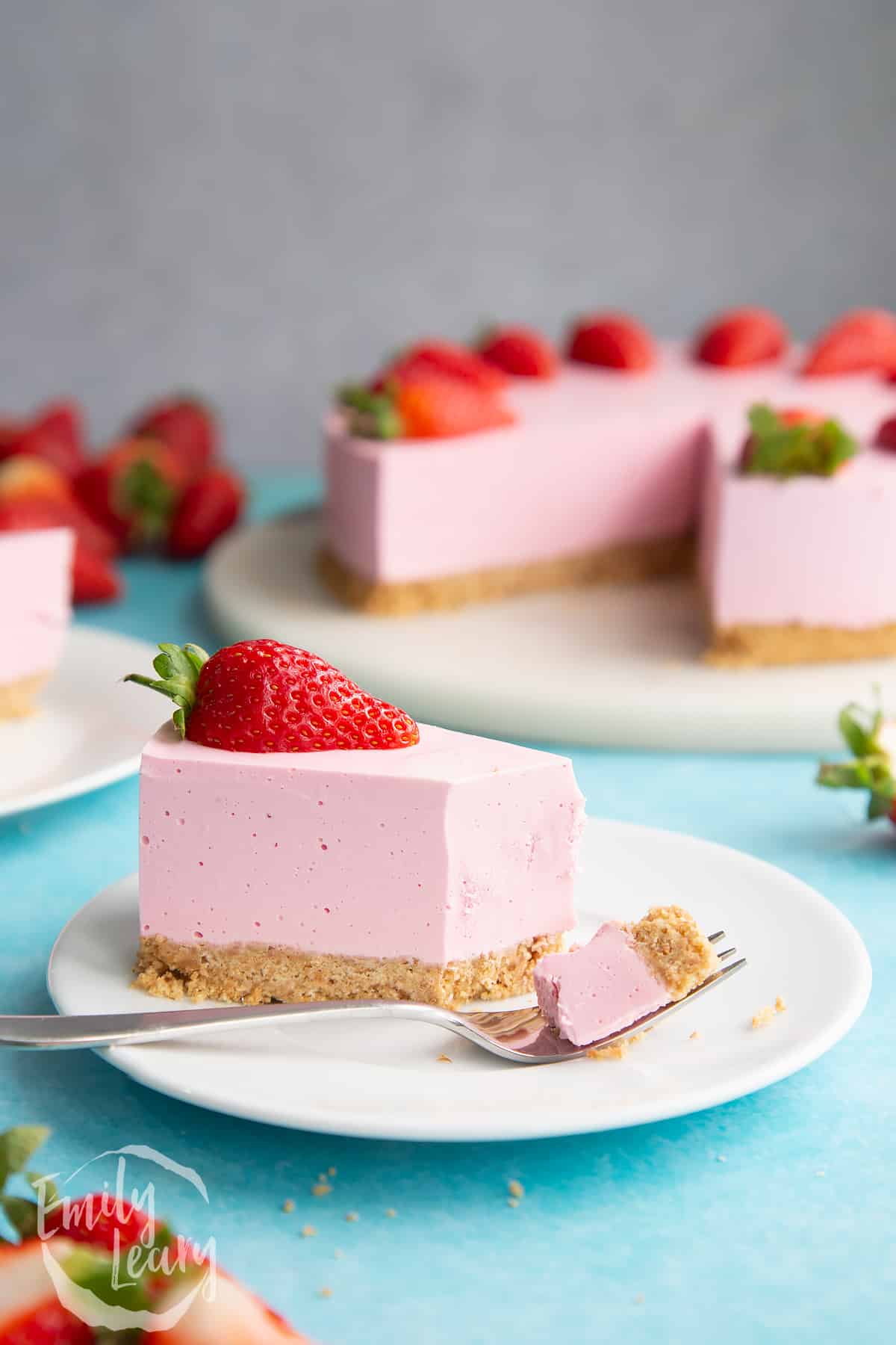 A slice of pink jello cheesecake on a white plate. It is topped with a strawberry half. Part has been eaten and some rests on a fork.