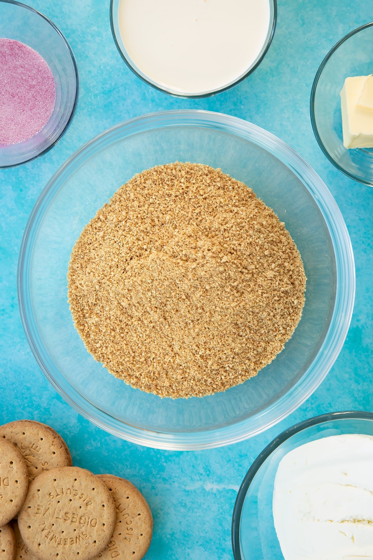 Crushed biscuits in a glass bowl. Ingredients to make jello cheesecake surround the bowl.