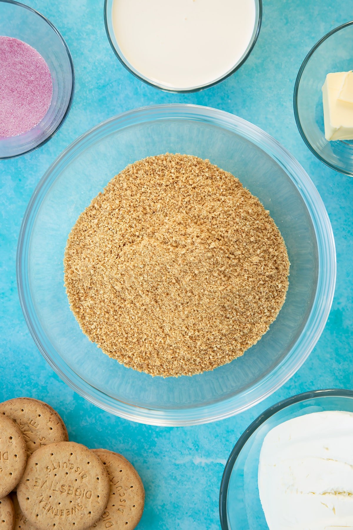 Crushed biscuits in a glass bowl. Ingredients to make jello cheesecake surround the bowl.