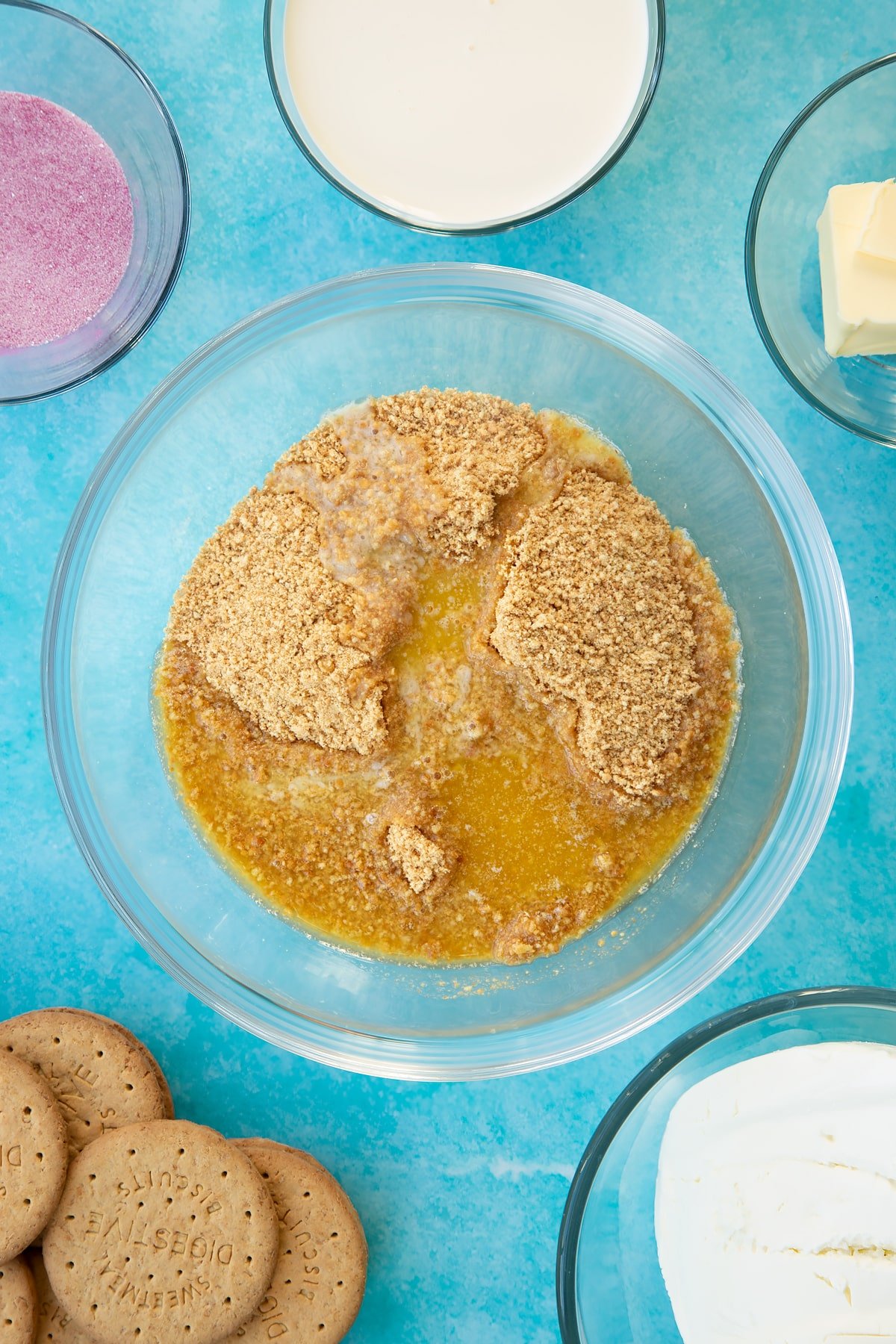 Crushed biscuits and melted butter in a glass bowl. Ingredients to make jello cheesecake surround the bowl.