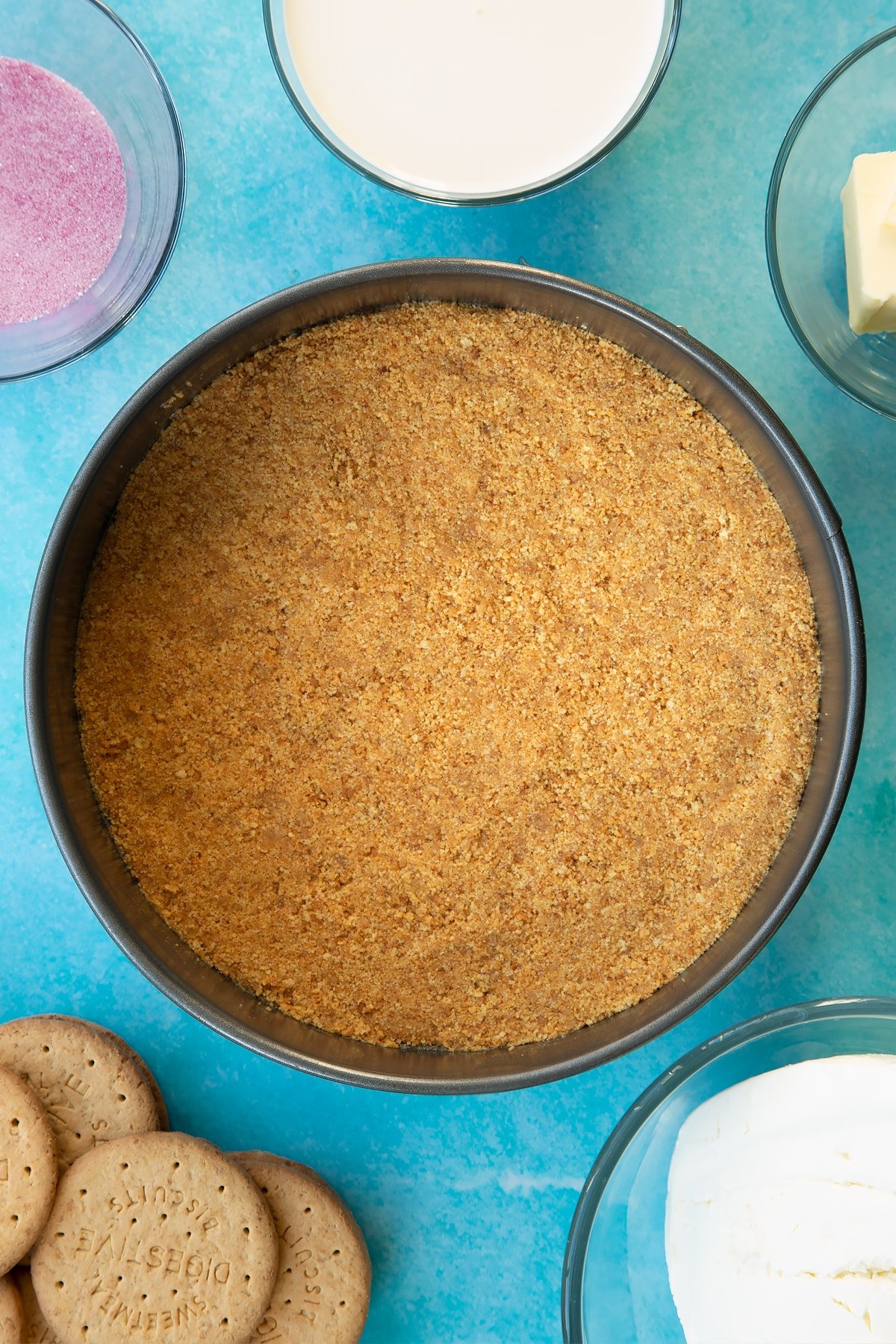 A biscuit base in a tin. Ingredients to make jello cheesecake surround the tin.