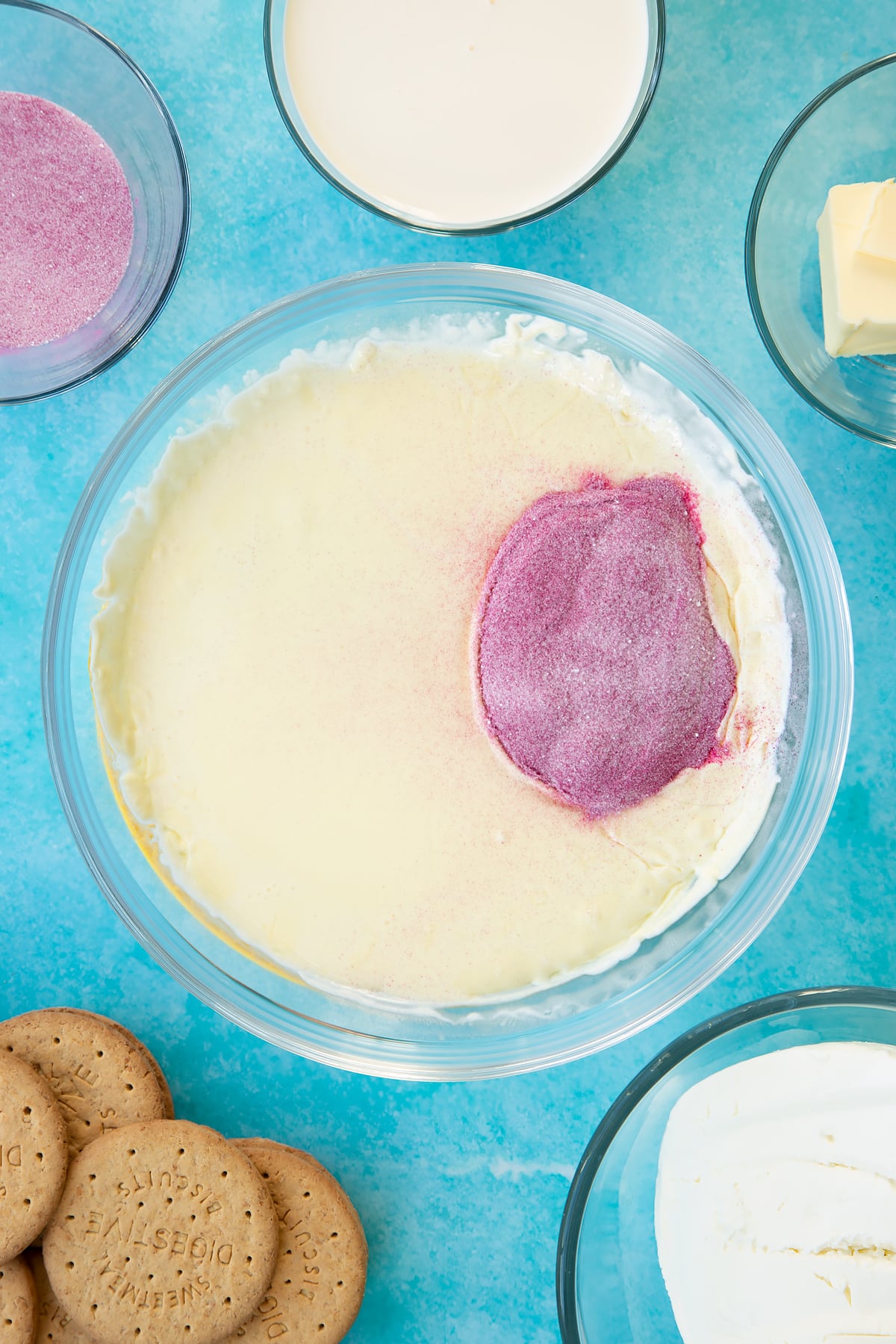 Hot double cream in a glass bowl with jello granules on top. Ingredients to make jello cheesecake surround the bowl.