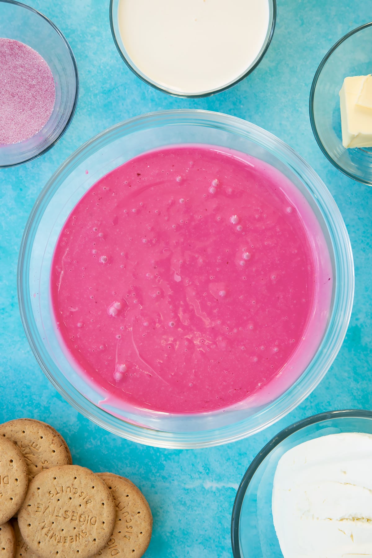 Hot double cream mixed with jello granules in a glass bowl. Ingredients to make jello cheesecake surround the bowl.