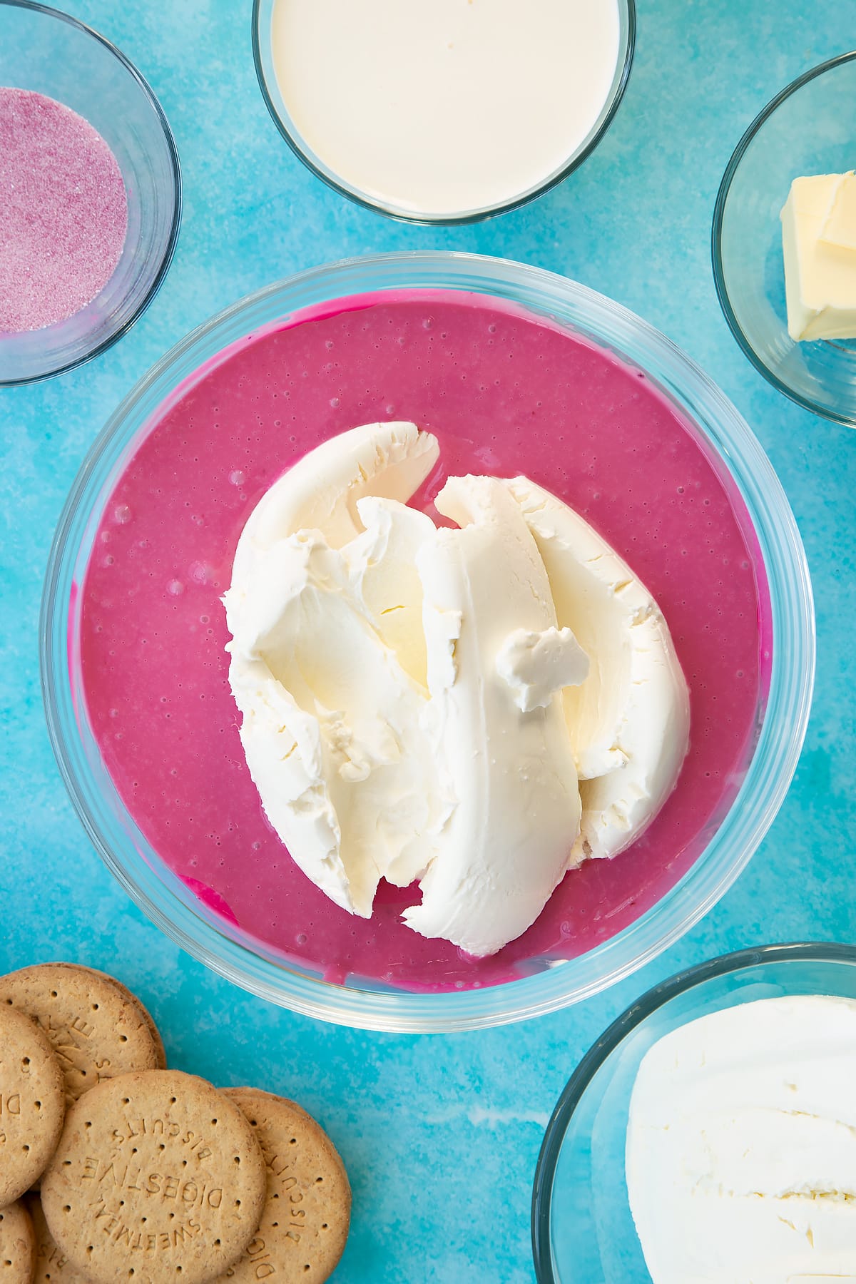 Hot double cream mixed with jello granules in a glass bowl with cream cheese on top. Ingredients to make jello cheesecake surround the bowl.