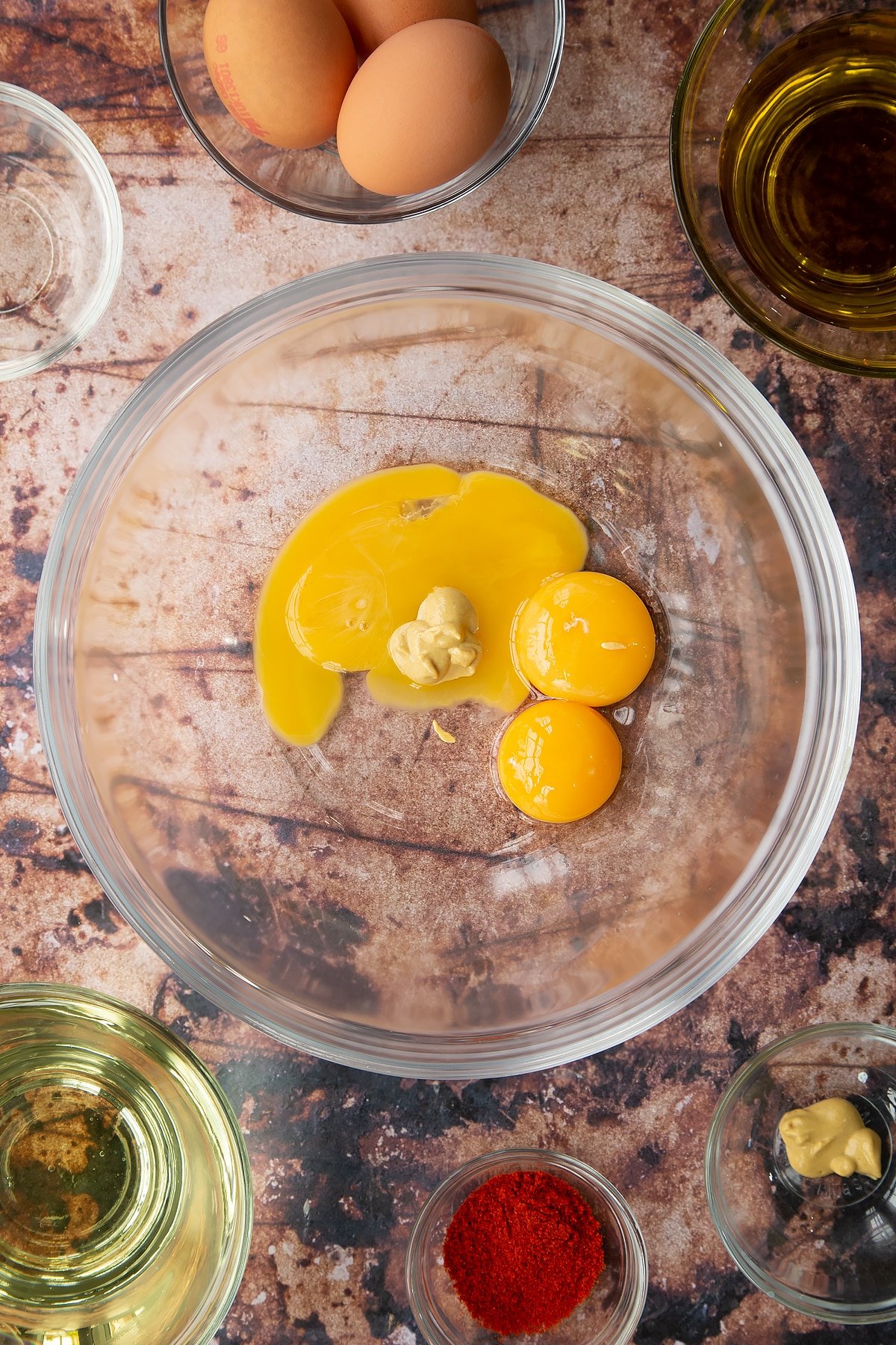 Vinegar, egg yolks and mustard in a glass mixing bowl. Ingredients to make paprika mayo surround the bowl.