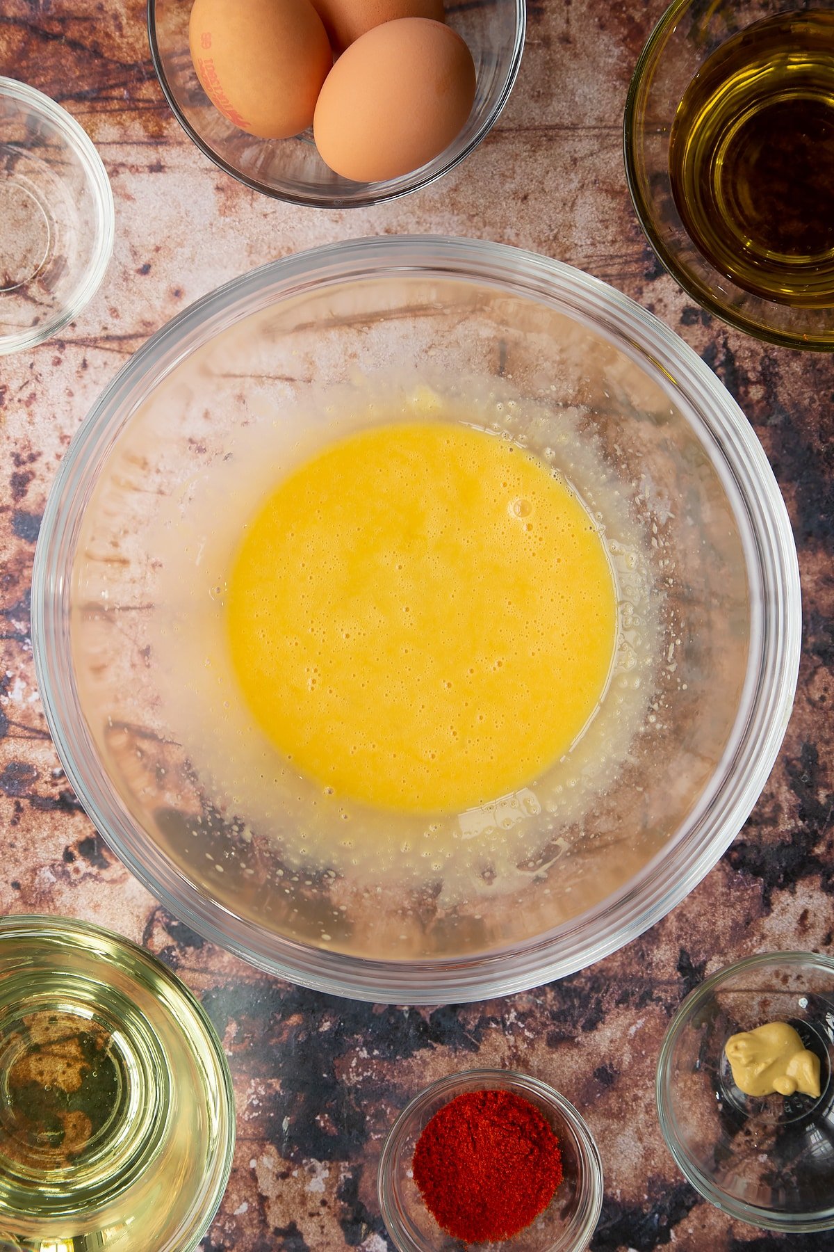 Vinegar, egg yolks and mustard whisked together in a glass mixing bowl. Ingredients to make paprika mayo surround the bowl.