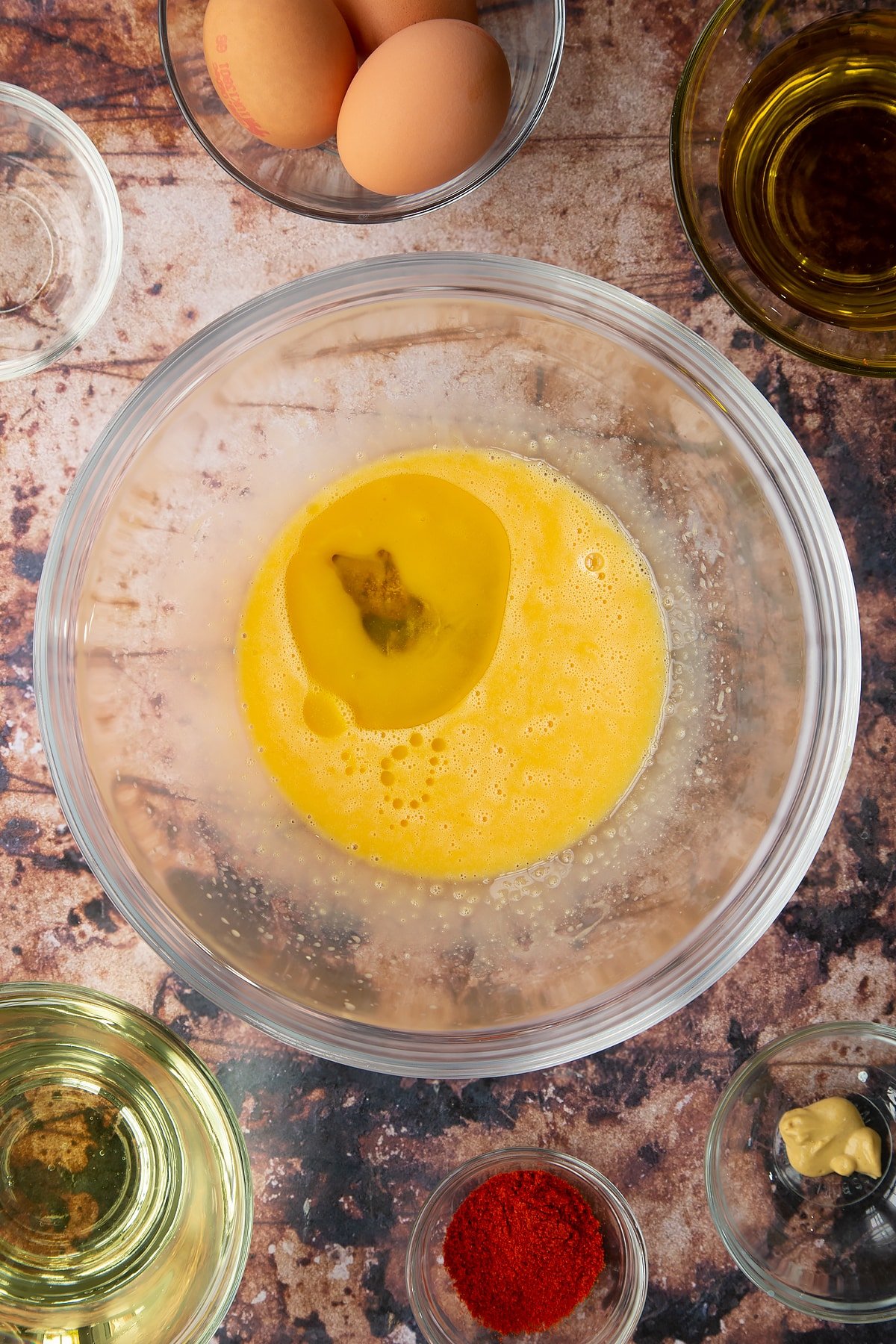 Vinegar, egg yolks and mustard whisked together in a glass mixing bowl with olive oil added. Ingredients to make paprika mayo surround the bowl.