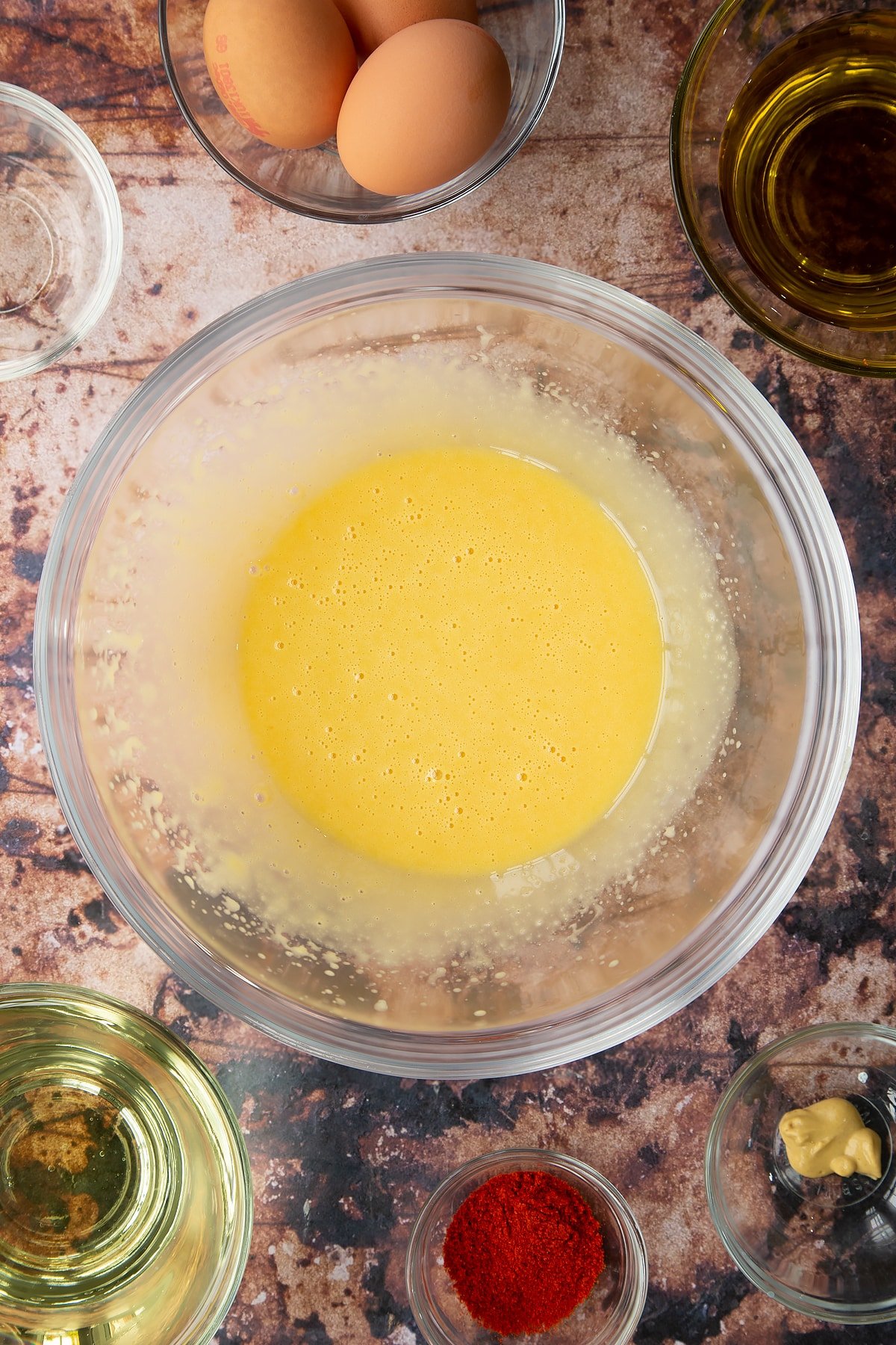 Vinegar, egg yolks and mustard whisked with oil in a glass mixing bowl. Ingredients to make paprika mayo surround the bowl.