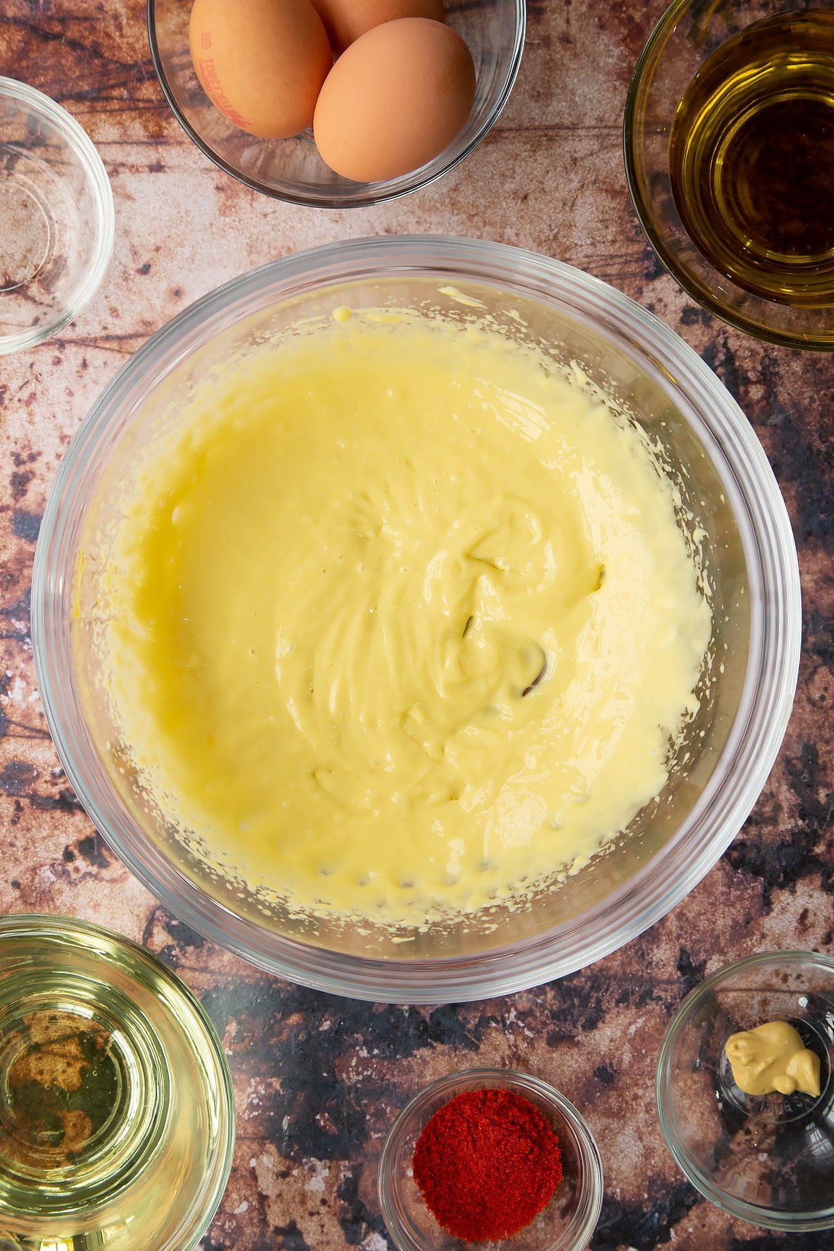 A small amount of thin mayo just beginning to come together in a  bowl. Ingredients to make paprika mayo surround the bowl.