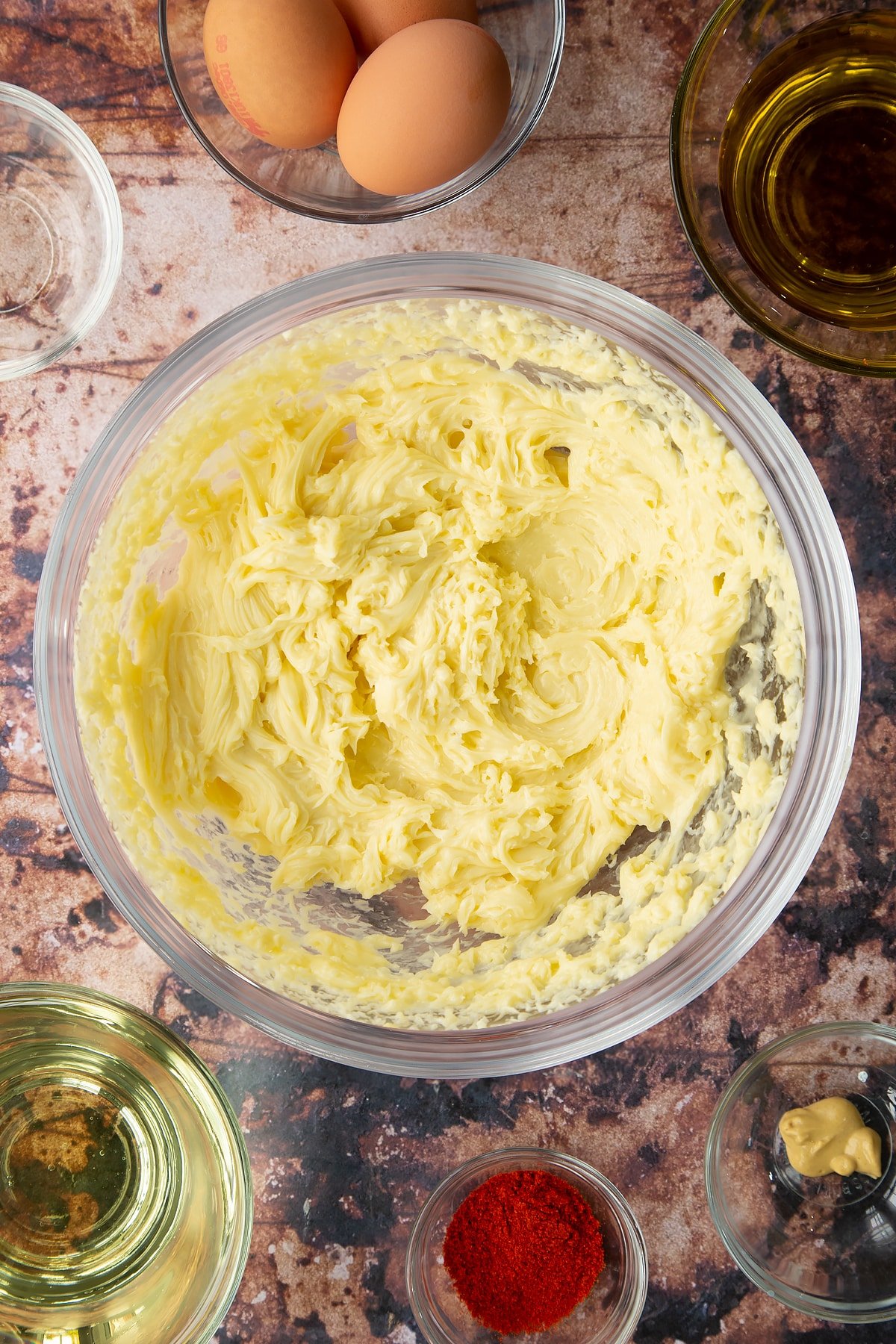 Thick olive oil mayo in a  bowl. Ingredients to make paprika mayo surround the bowl.