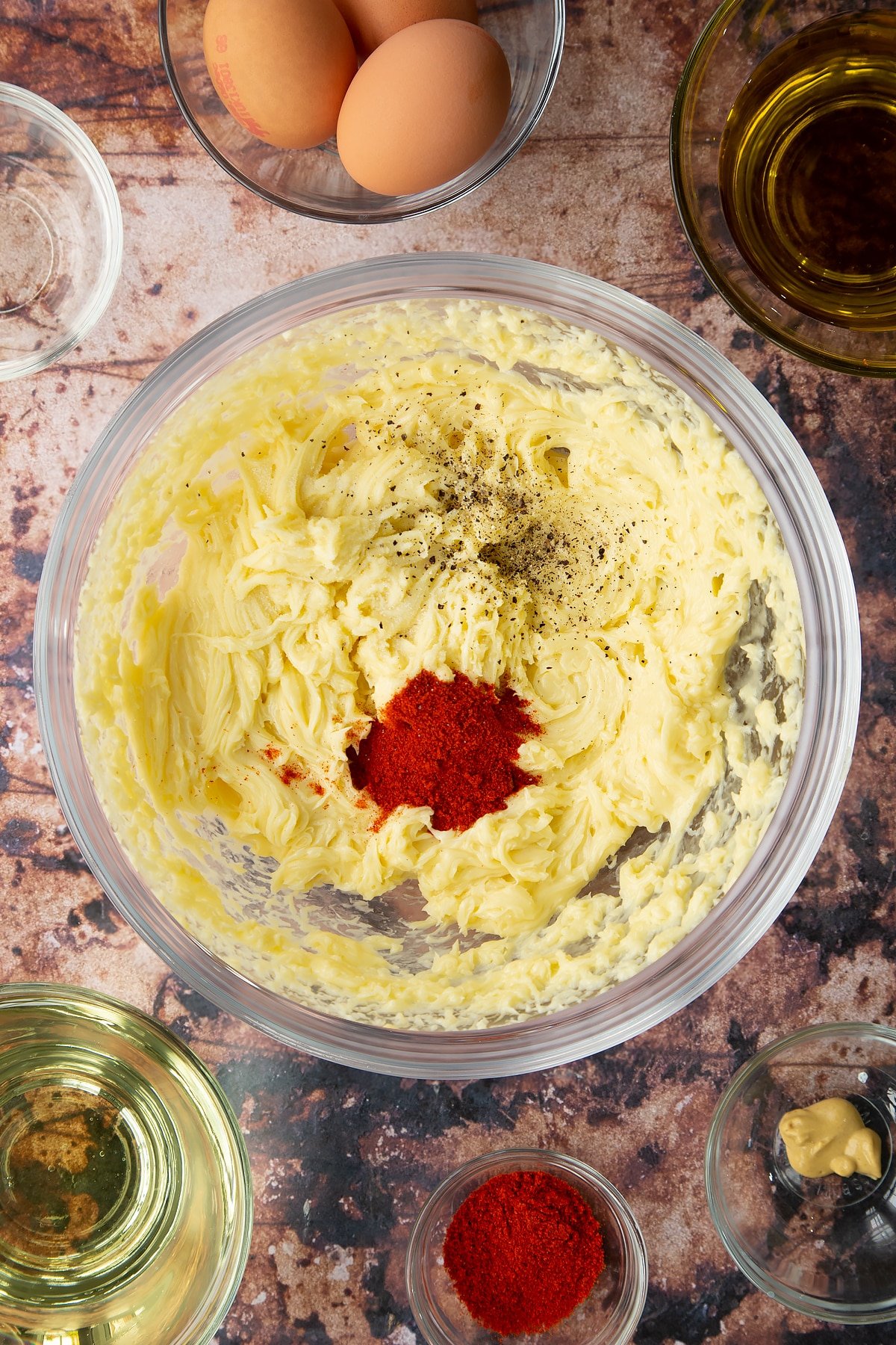 Thick olive oil mayo in a  bowl with paprika and pepper on top. Ingredients to make paprika mayo surround the bowl.