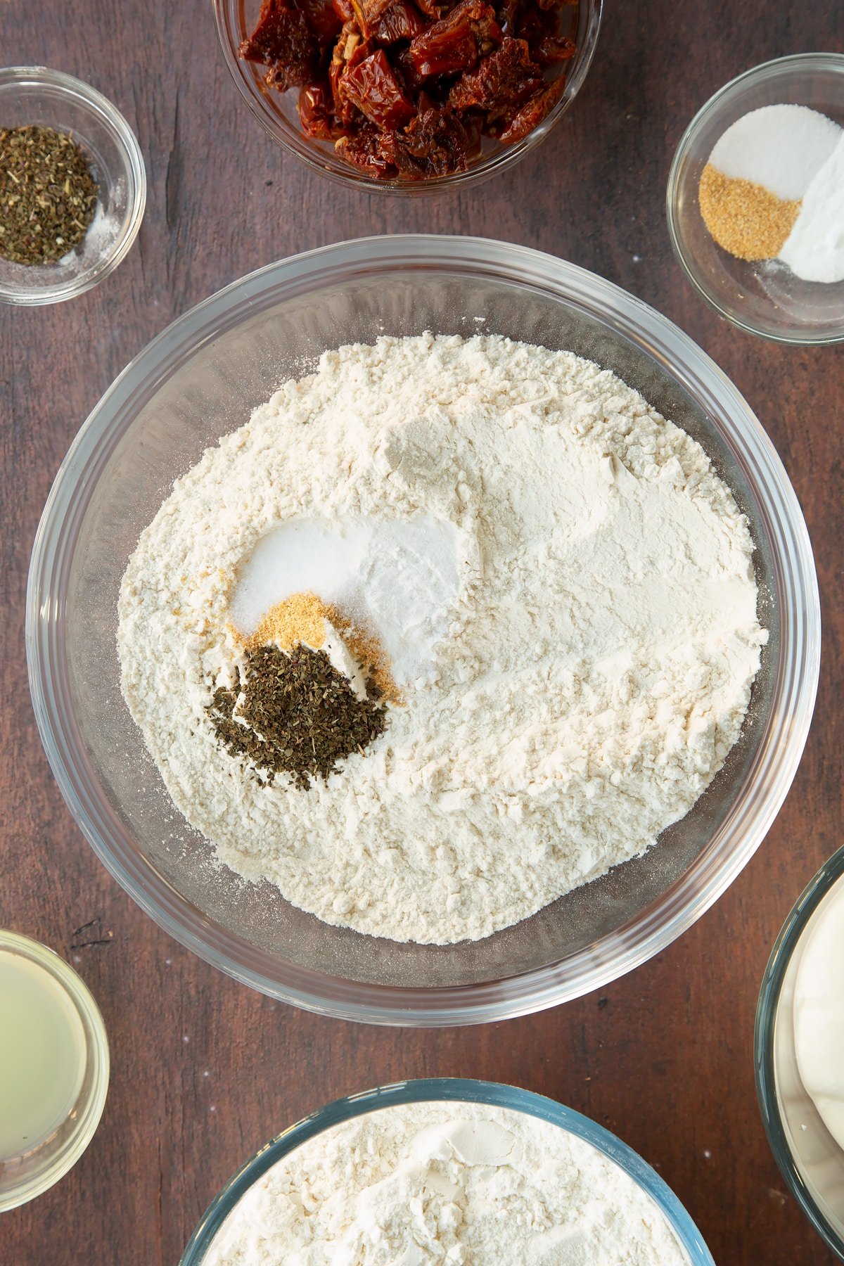 Flour, salt, bicarbonate of soda, garlic granules, dried basil and a pinch of salt and pepper in a bowl. Ingredients to make Sundried tomato soda bread surround the bowl.