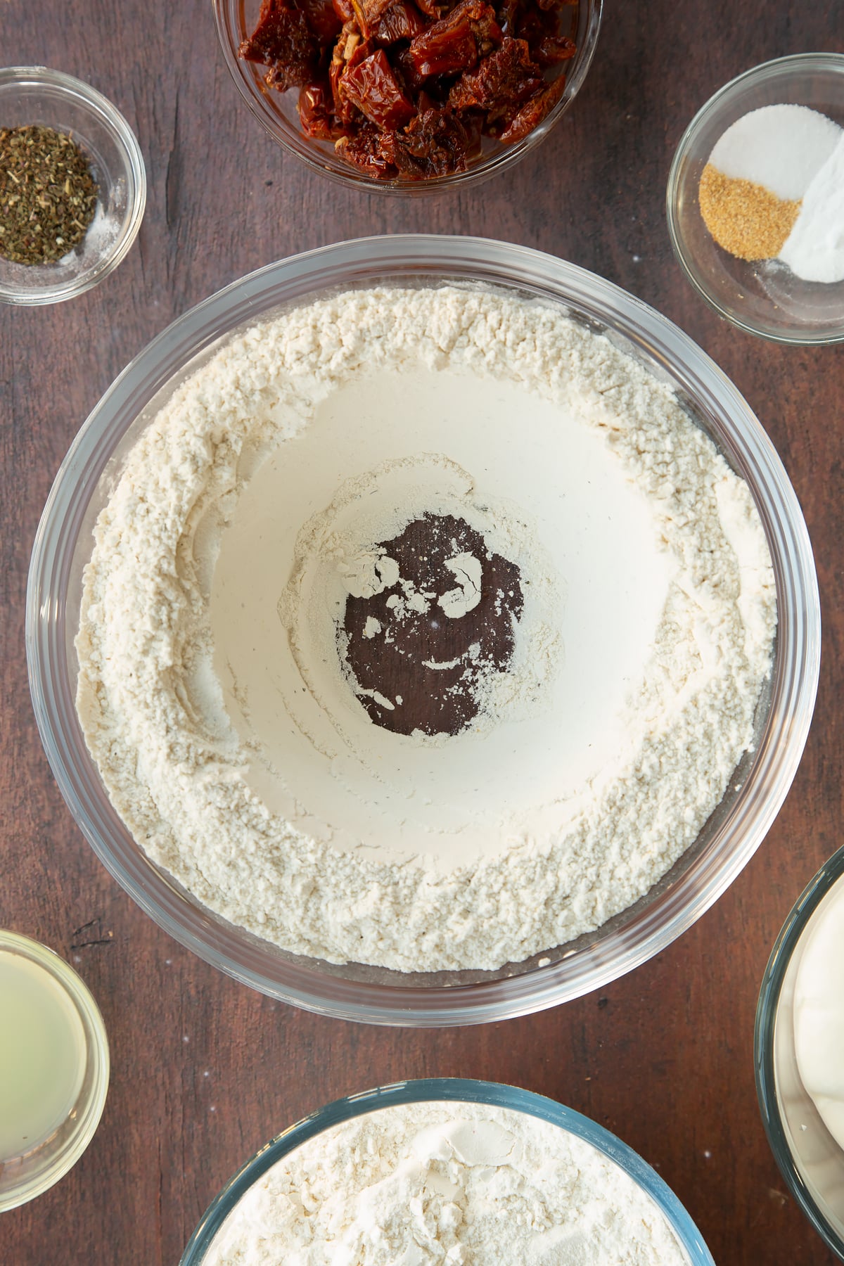 Flour, bicarbonate of soda and spices in a bowl with a well in the middle. Ingredients to make Sundried tomato soda bread surround the bowl.